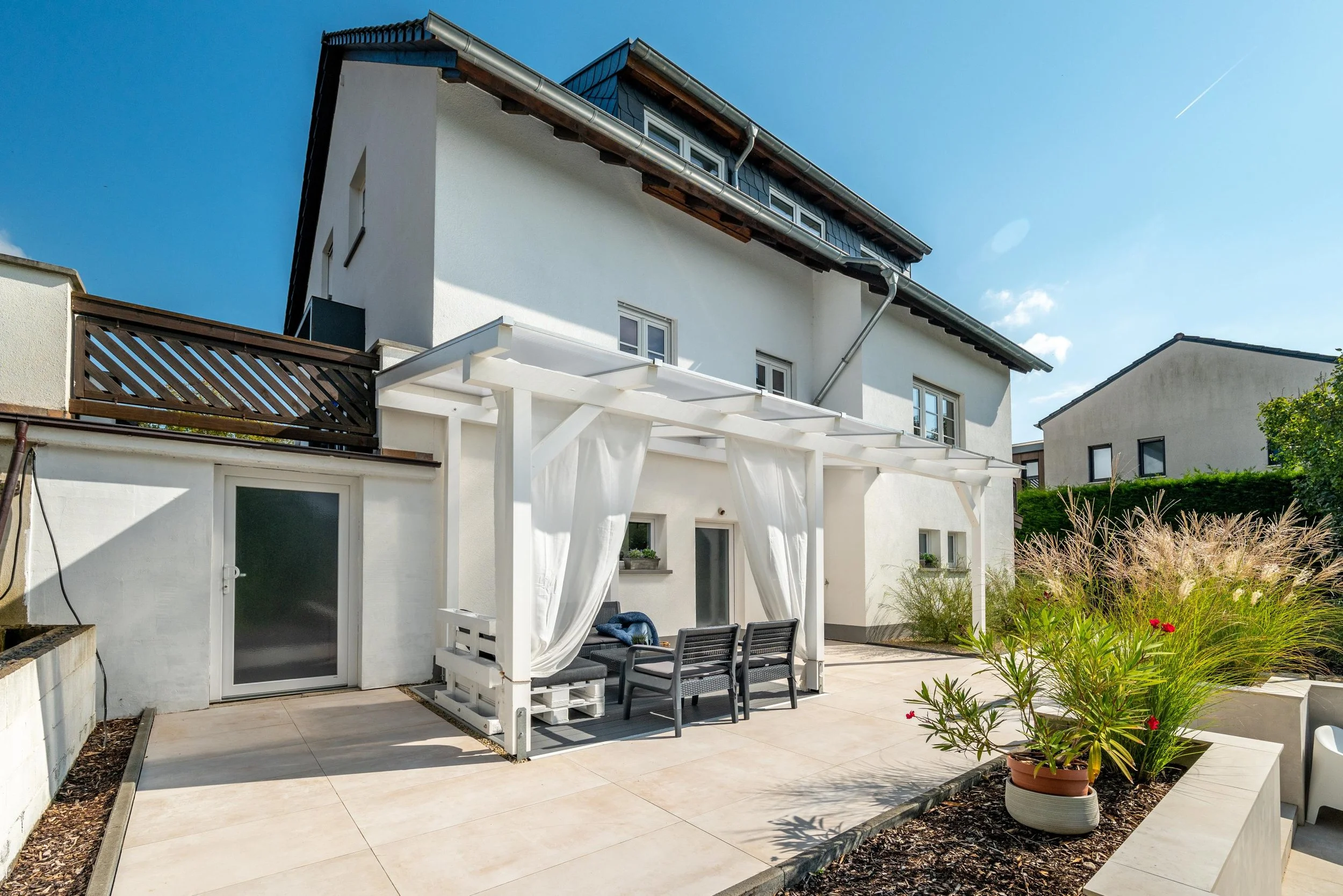Le patio arrière d'une maison blanche avec une pergola avec des rideaux, un mobilier lounge, et des plantes en pot