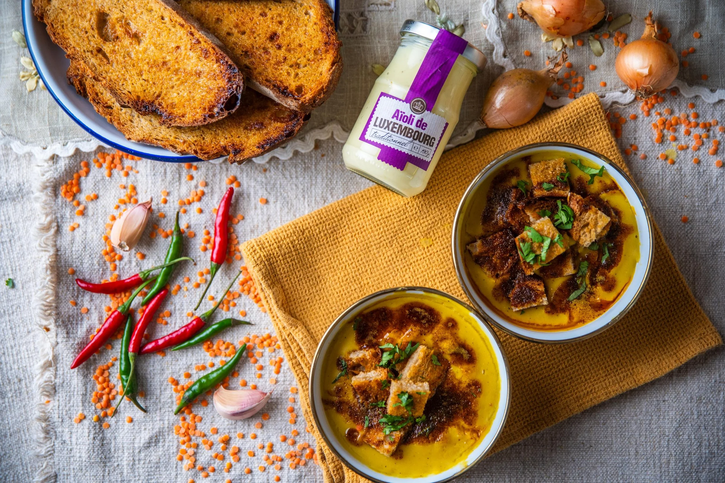 Deux bols de soupe de poisson avec des morceaux de poisson, garnis de persil, sur un napperon en lin, accompagnés de pain grillé, d'oignons, de carottes, d'ail, de piment et d'une jarre d'aïoli de Luxembourg.