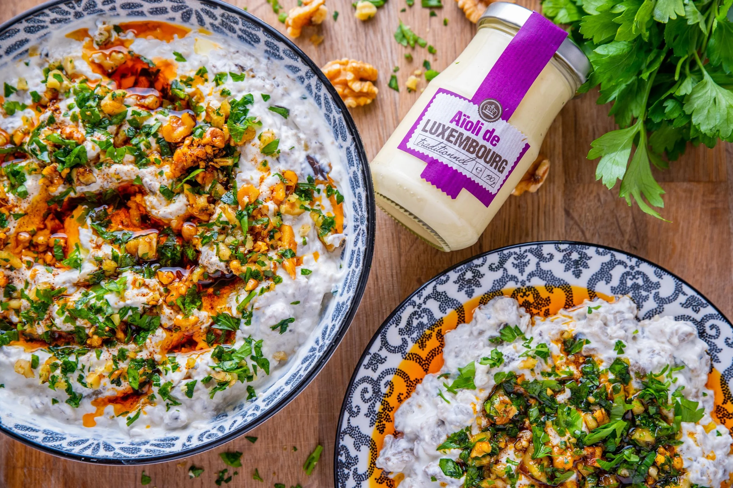 Deux plats de dip à la crème avec des herbes, des noix et de l'huile pimentée, accompagnés d'un pot de crème d'ail du Luxembourg, décorés avec du persil, sur une table en bois.