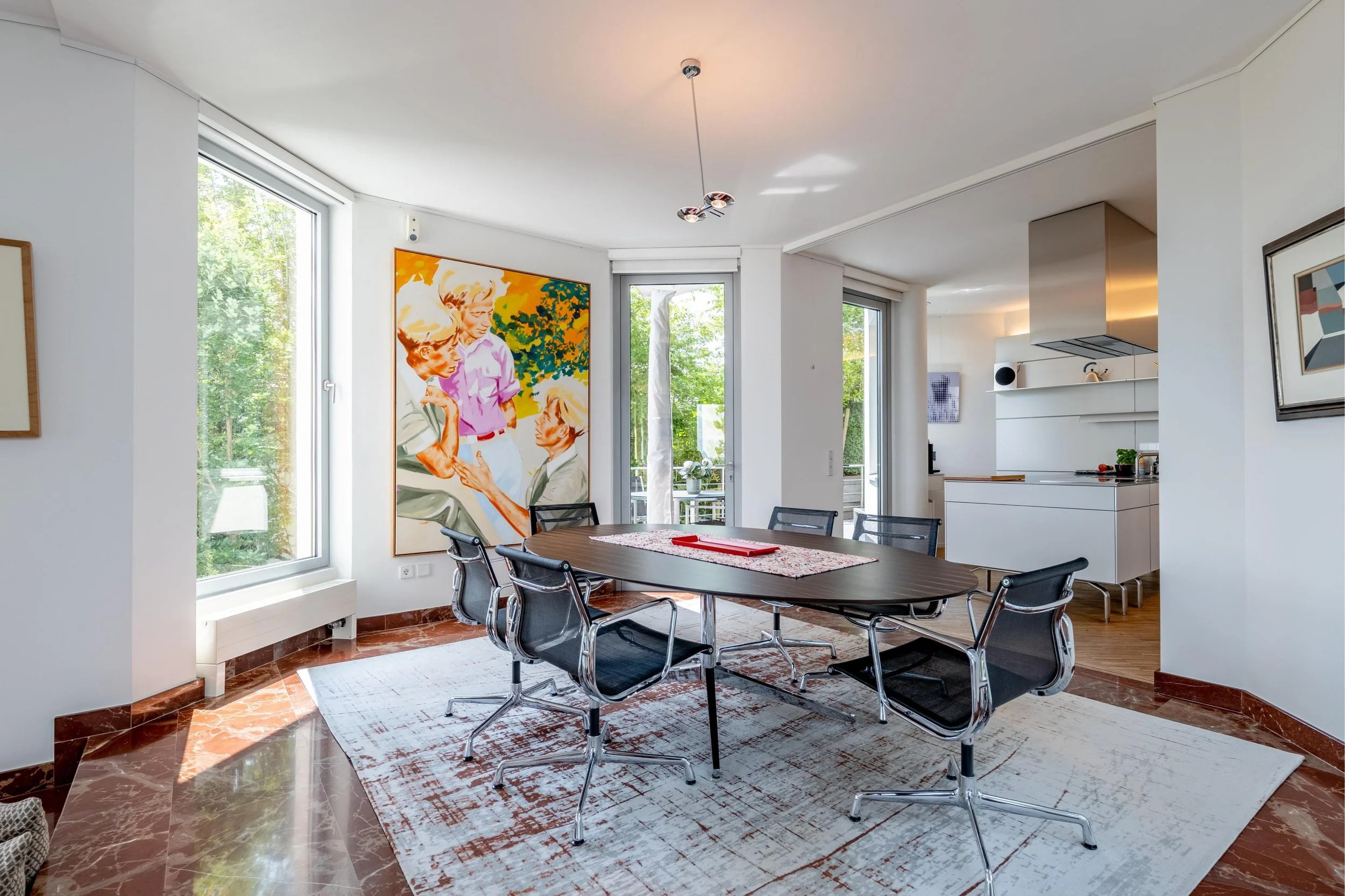 Salle à manger moderne avec un tableau coloré représentant plusieurs personnes, un tapis blanc, une table ovale en bois avec six chaises noires et un lustre sculptural suspendu, cuisine blanche en arrière-plan, grandes fenêtres laissant entrer la lum