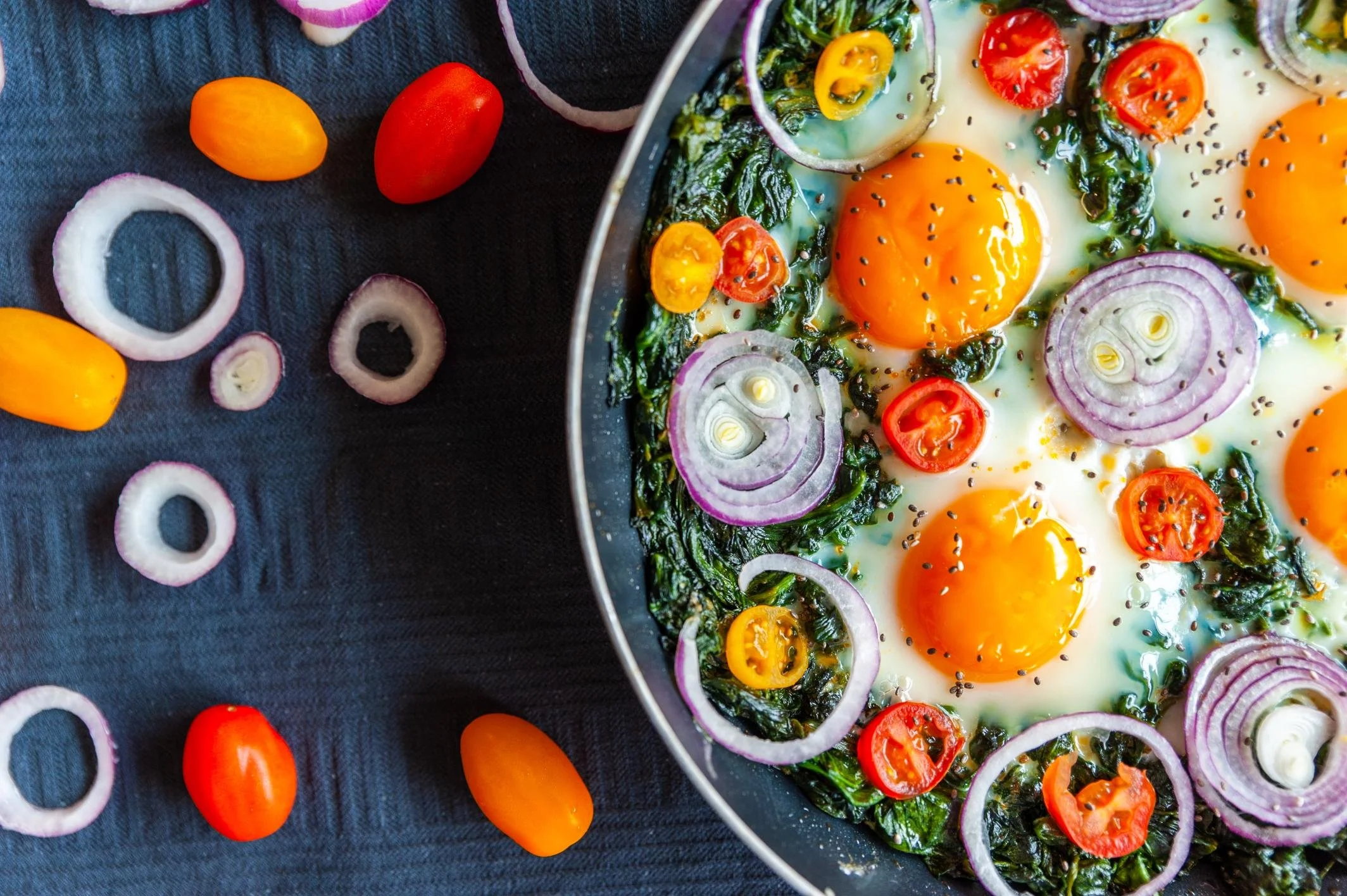 Oeufs au plat cuits avec des tomates cerises coupées, des rondelles d'oignon et des épinards dans une poêle, avec quelques tomates cerises et rondelles d'oignon disposées autour sur une nappe noire.