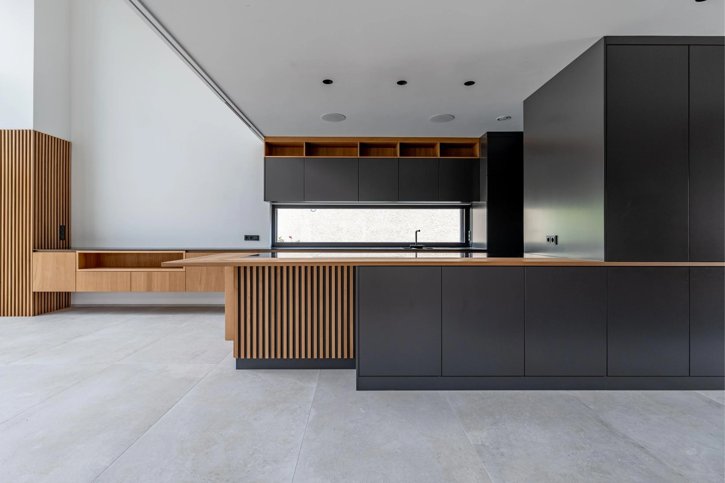 Cuisine moderne avec éléments en bois naturel et armoires noires, grande fenêtre, sol en carrelage clair.