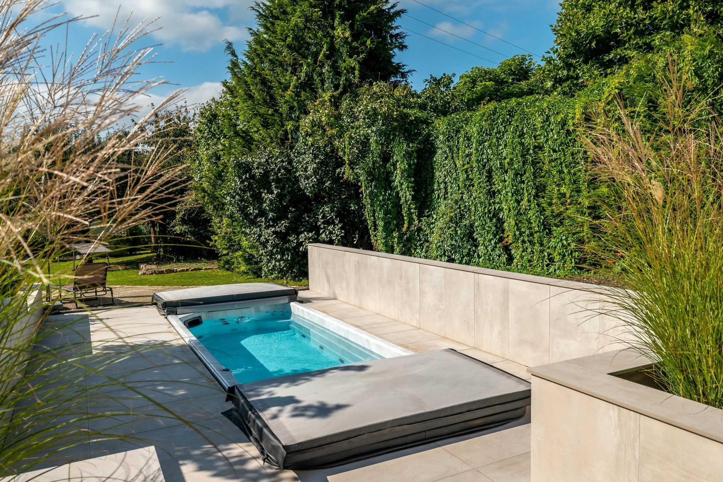 Une piscine extérieure dans un jardin verdoyant avec des arbres et des arbustes, sous un ciel bleu avec quelques nuages.