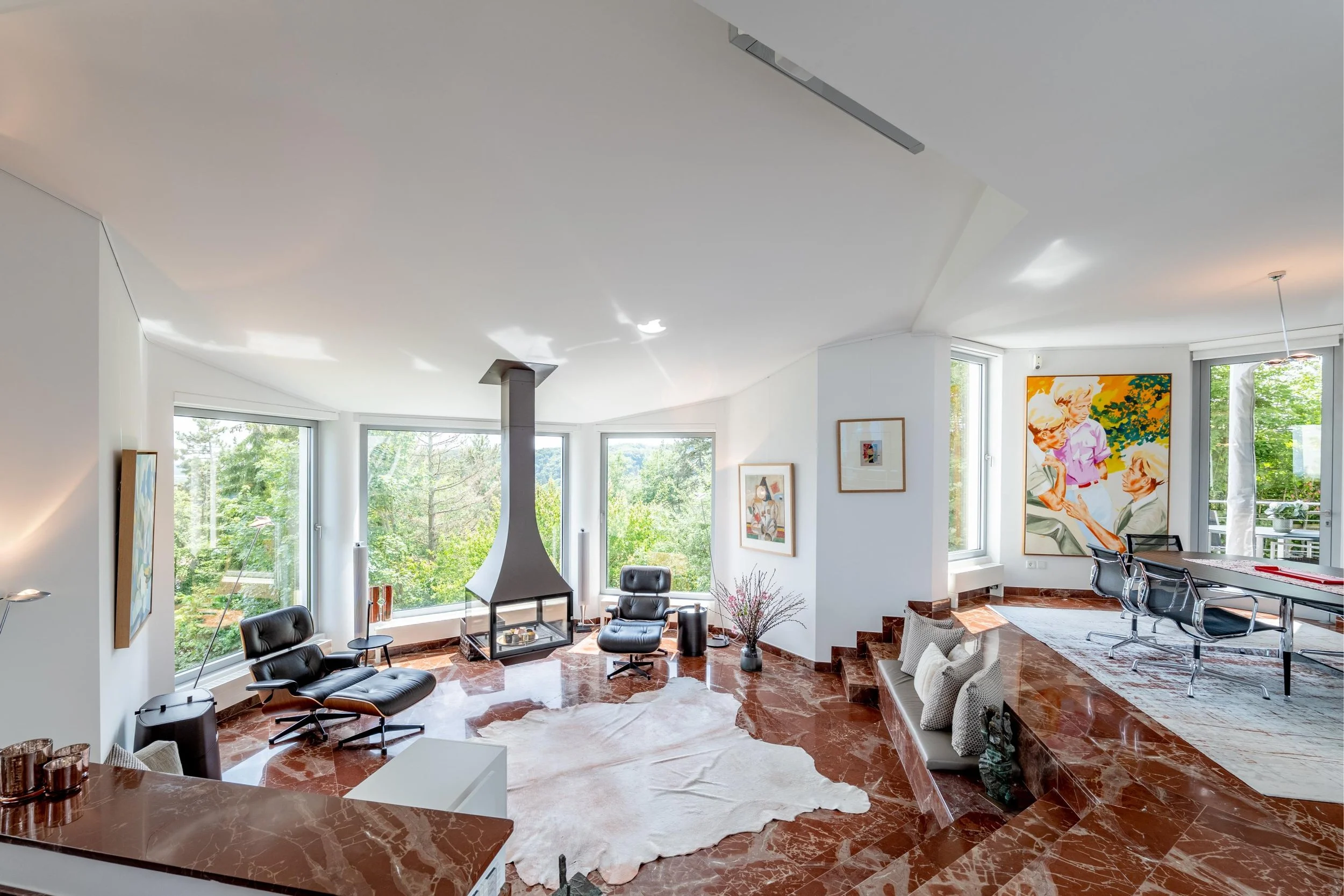 Salon moderne avec cheminée en verre, deux fauteuils en cuir noir, une grande fenêtre offrant une vue sur la nature, un tapis blanc, des œuvres d'art sur les murs, et un espace de réunion avec table et chaises.
