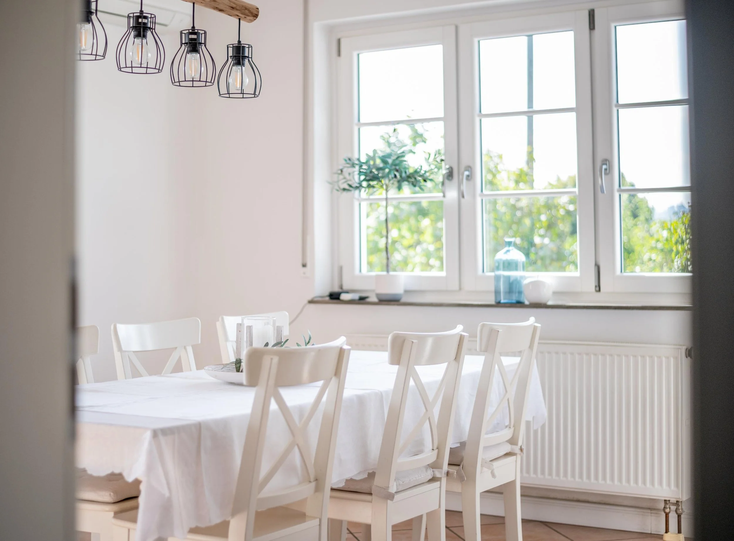 Salle à manger lumineuse avec une table rectangulaire recouverte d'une nappe blanche, entourée de six chaises blanches. Une grande fenêtre laisse entrer la lumière naturelle, avec des vases décoratifs sur le rebord de la fenêtre et un arbre à l'intér