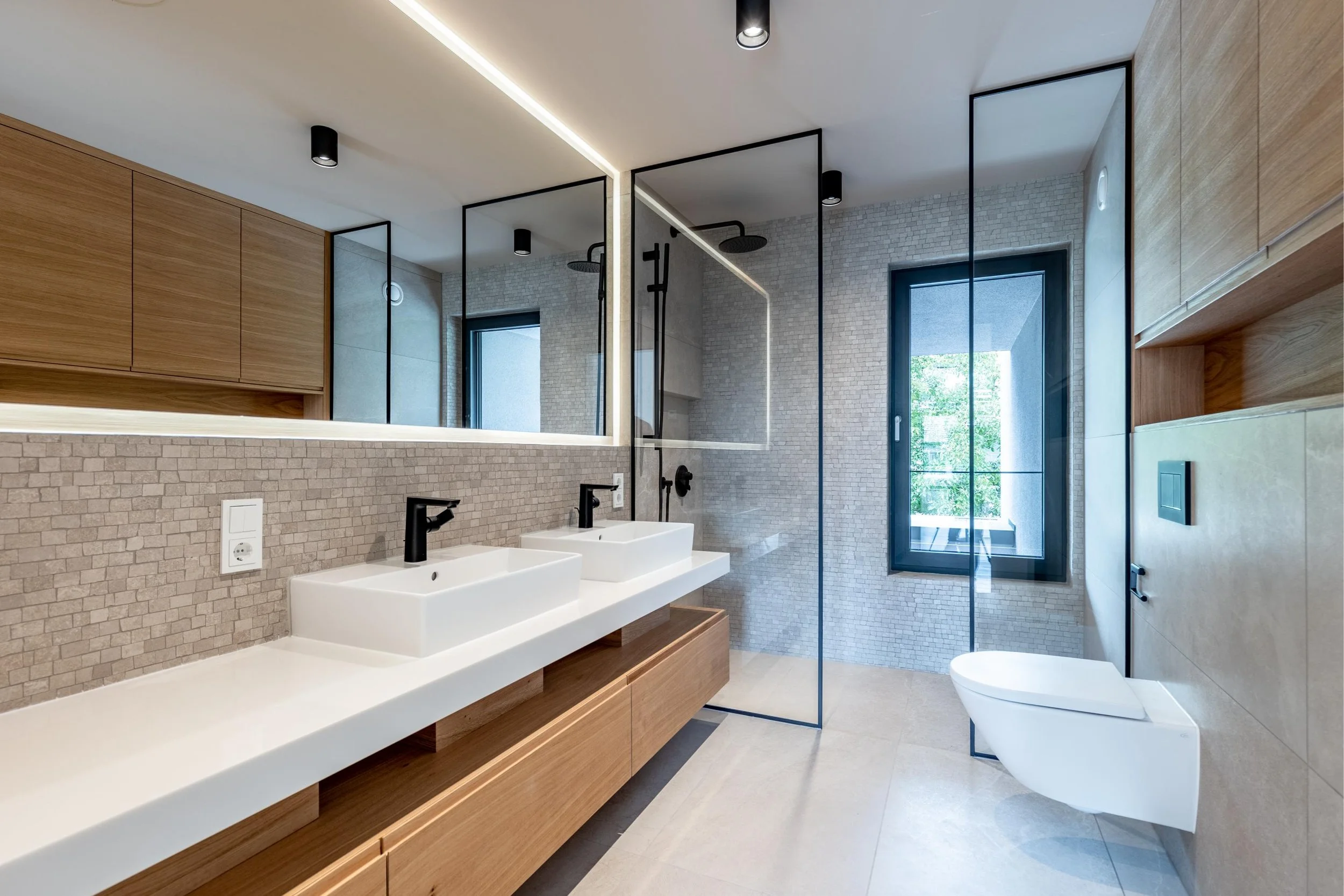 Salle de bain moderne avec deux lavabos, grand miroir, cabine de douche avec parois en verre, toilettes suspendues et fenêtre laissant entrer de la lumière naturelle.