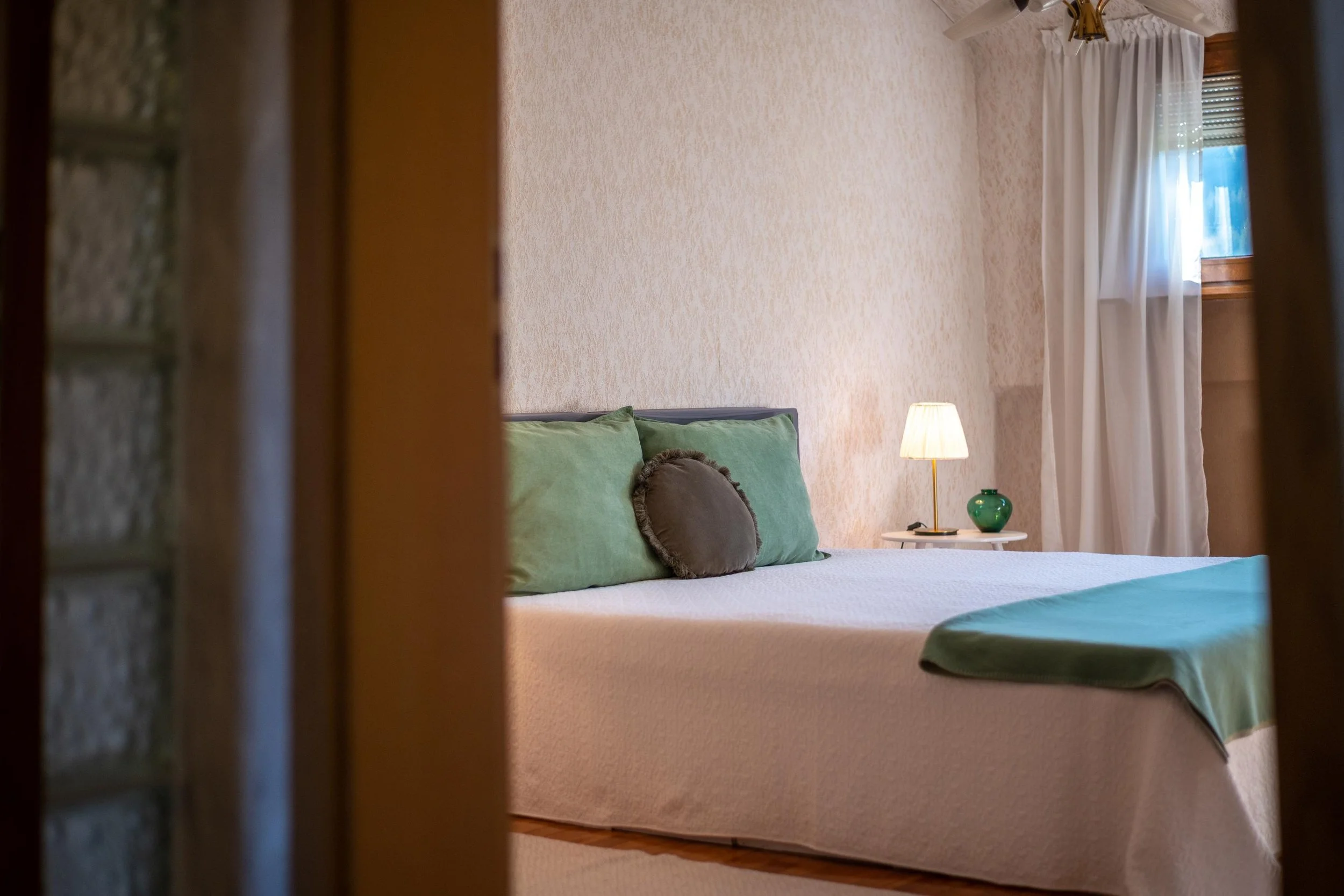 Chambre avec un lit à trois coussins verts et un coussin rond, une table de nuit avec une lampe, une fenêtre avec rideaux blancs.