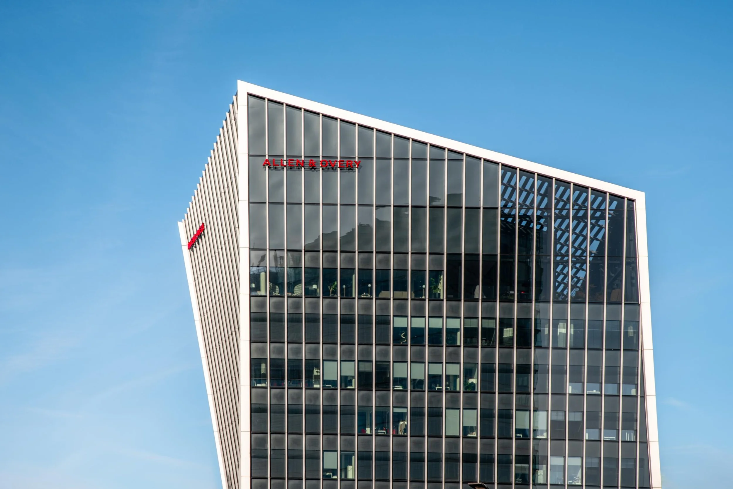 Gratte-ciel moderne avec des fenêtres en verre, portant le nom 'ALLEN & OVERY' en lettres rouges, contre un ciel bleu.