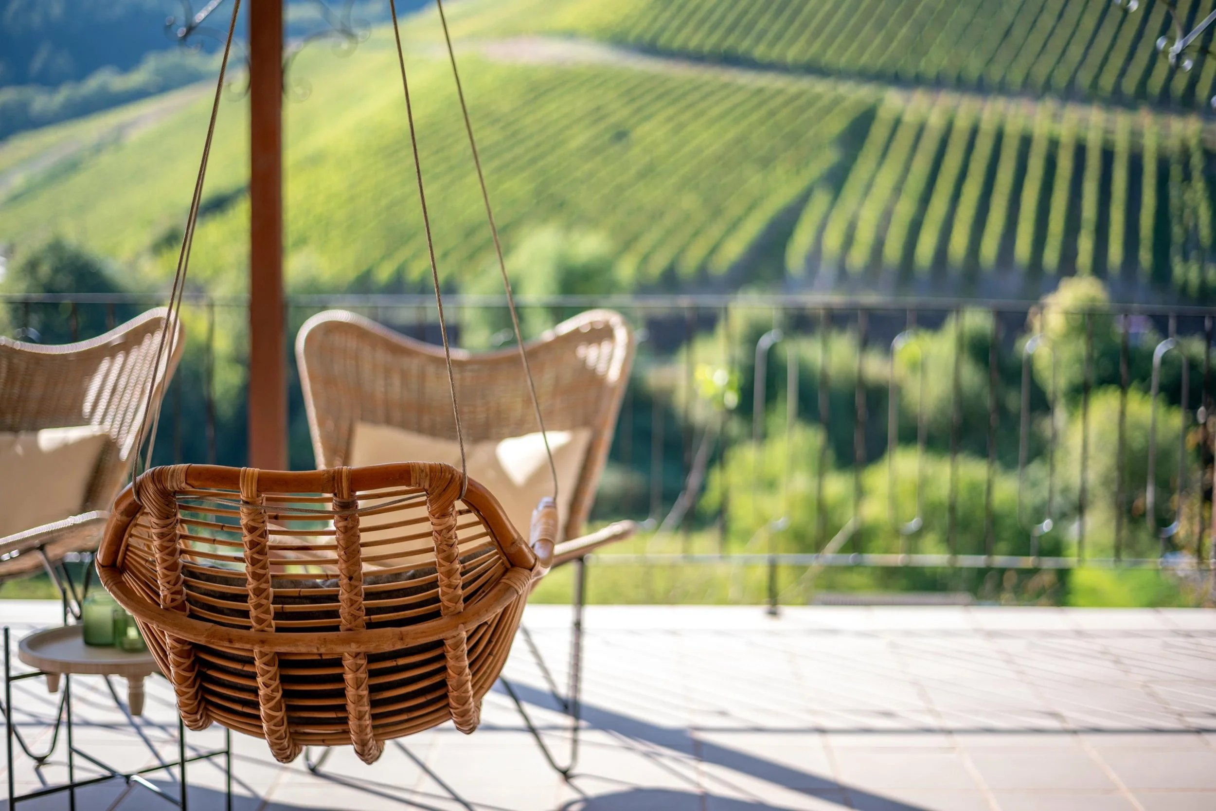 Une balançoire en rotin suspendue sur une terrasse en bois, avec deux chaises en rotin en arrière-plan, surplombant un paysage de vignes verdoyantes sur une colline ensoleillée.