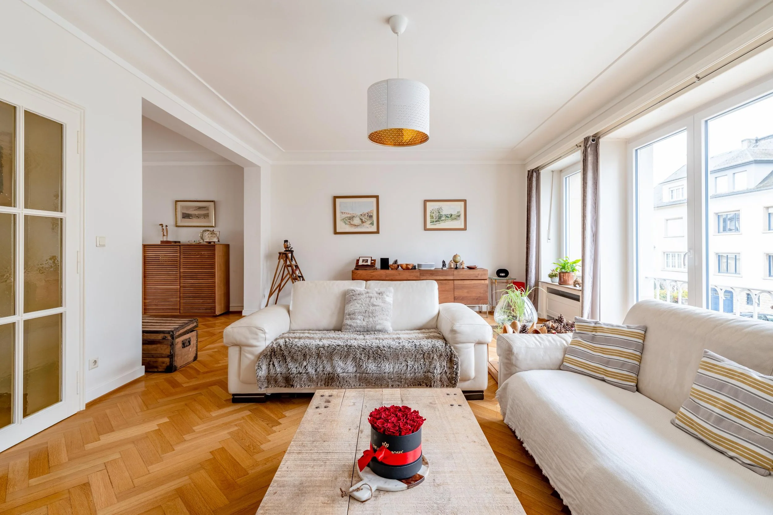 Salon lumineux avec deux canapés blancs, un tapis en fausse fourrure, une table en bois avec un bouquet de roses rouges, et de grandes fenêtres laissant entrer la lumière.