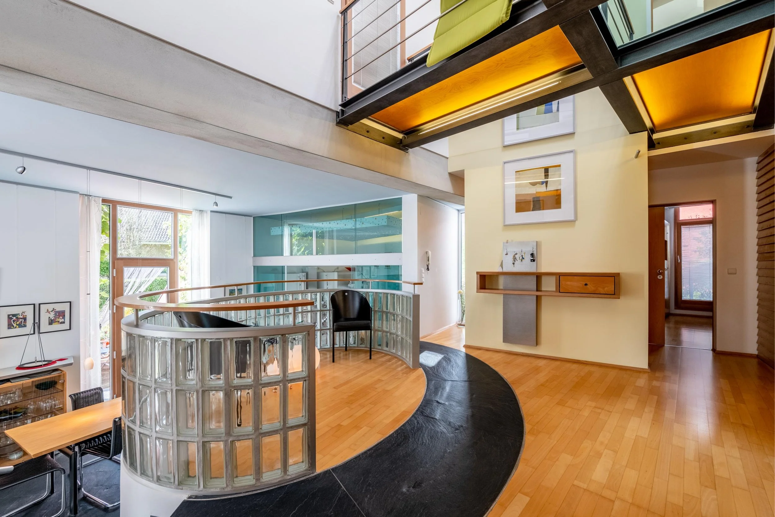 Intérieur moderne d'une maison avec un plancher en bois clair, un mur jaune, des cadres muraux, une mezzanine avec une balustrade en verre, un fauteuil noir, une porte-fenêtre et un mur en verre.