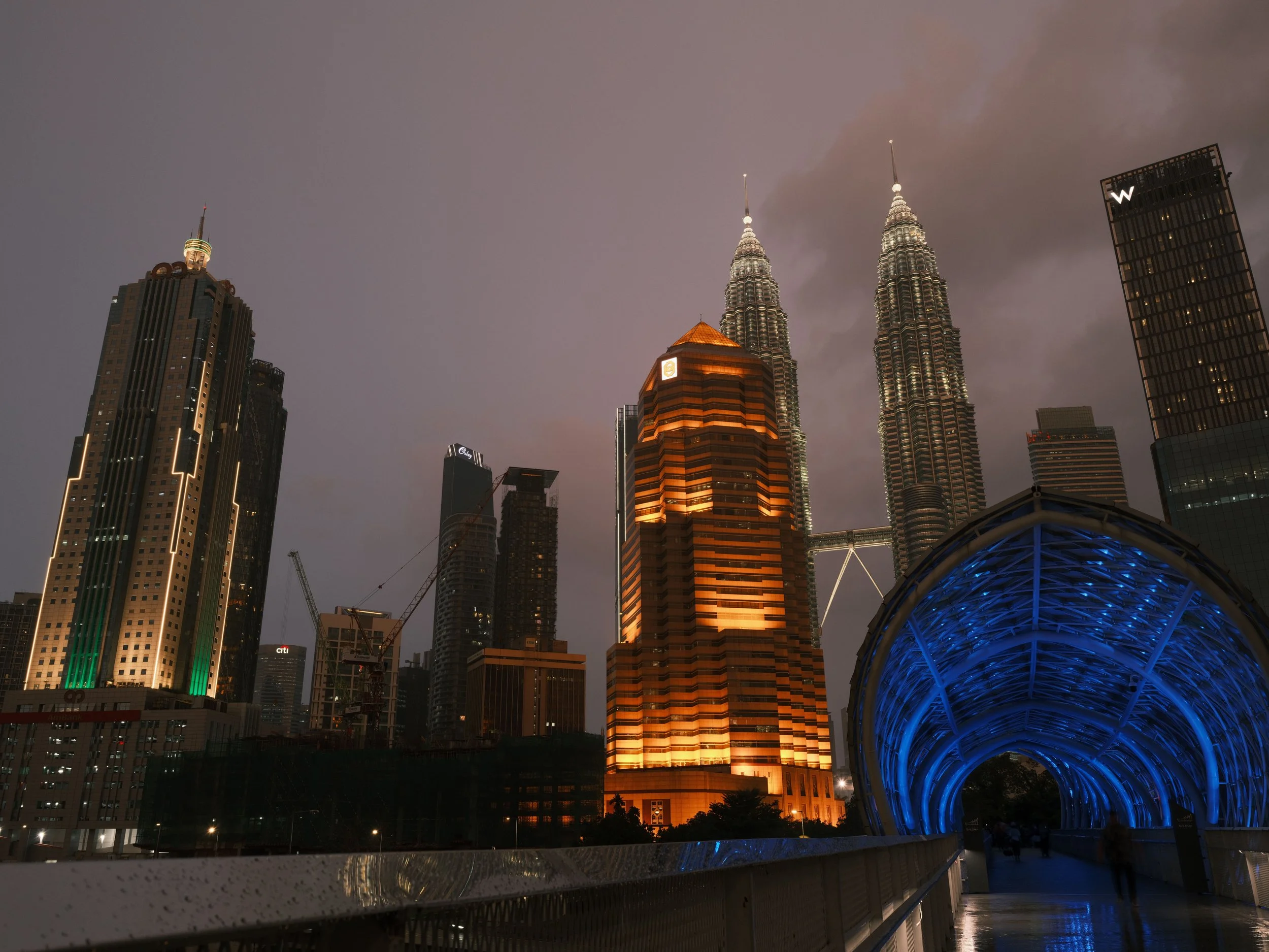 Nighttime view of Kuala Lumpur skyline featuring the illuminated Petronas Twin Towers and surrounding skyscrapers, with a futuristic blue-lit pedestrian bridge in the foreground.
