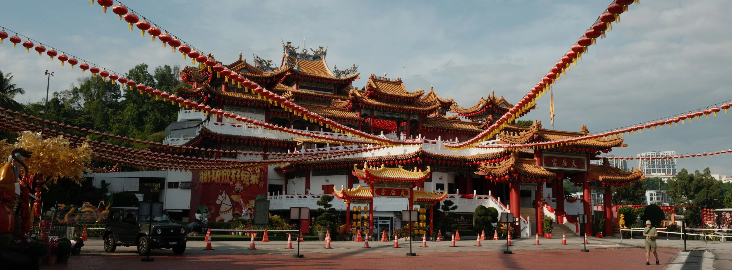 Traditional Chinese temple decorated with red lanterns, ornate roofing, and intricate architectural details, with a hillside and modern buildings in the background.