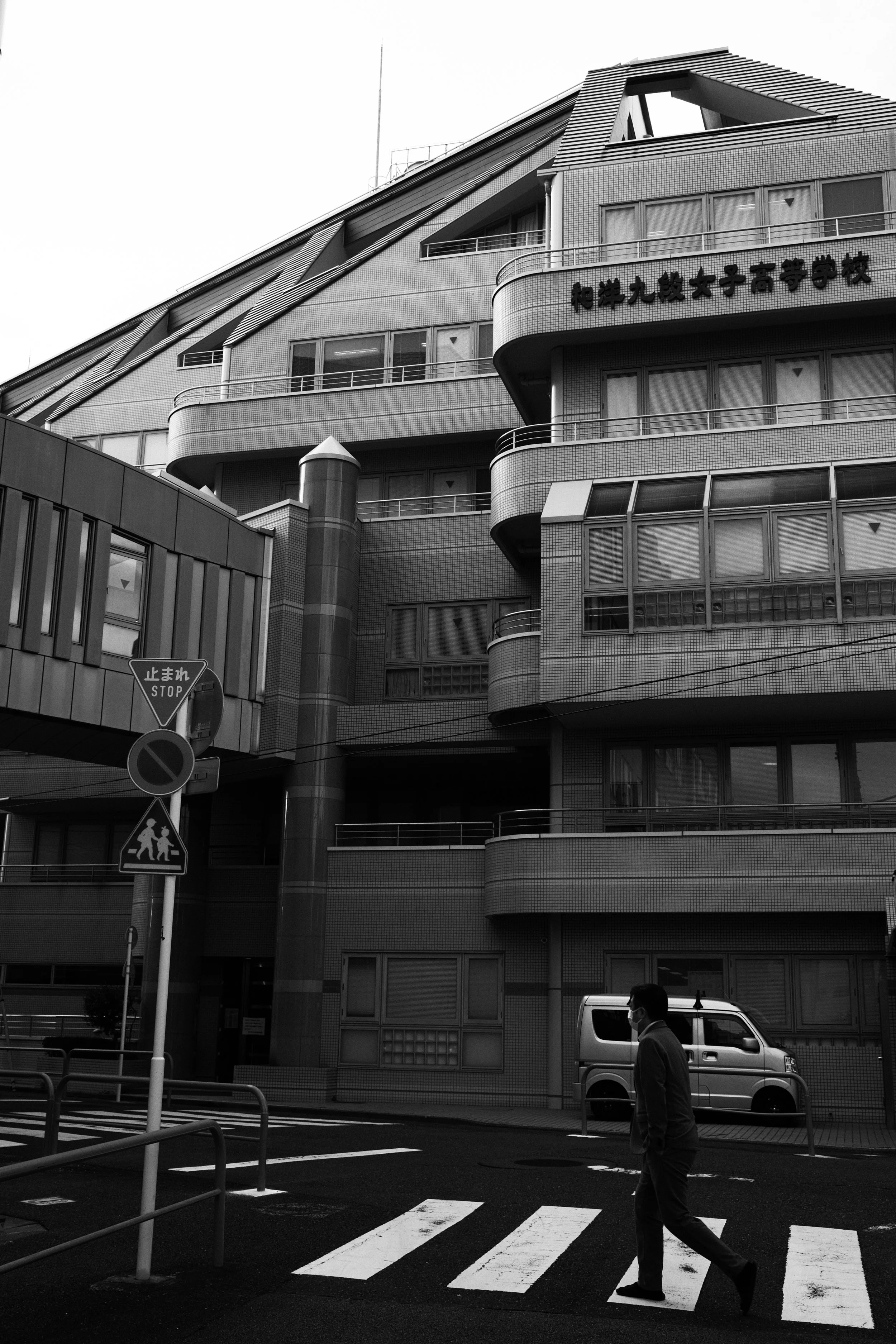 Black and white photo of a city street scene with a high-rise building, a pedestrian crossing, and a man wearing a face mask walking across the crosswalk.