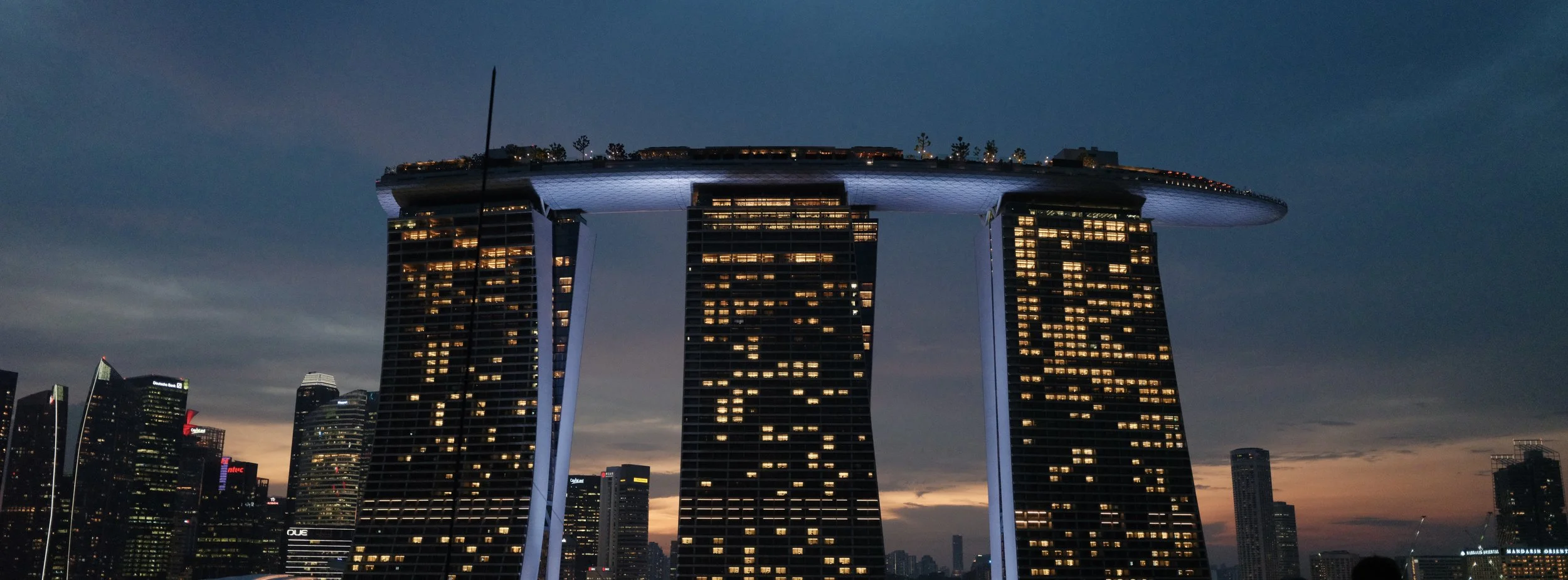 Nighttime view of the Marina Bay Sands hotel in Singapore, with three tall towers connected by a large rooftop structure, illuminated with lights against a dark skyline and cloudy sky.