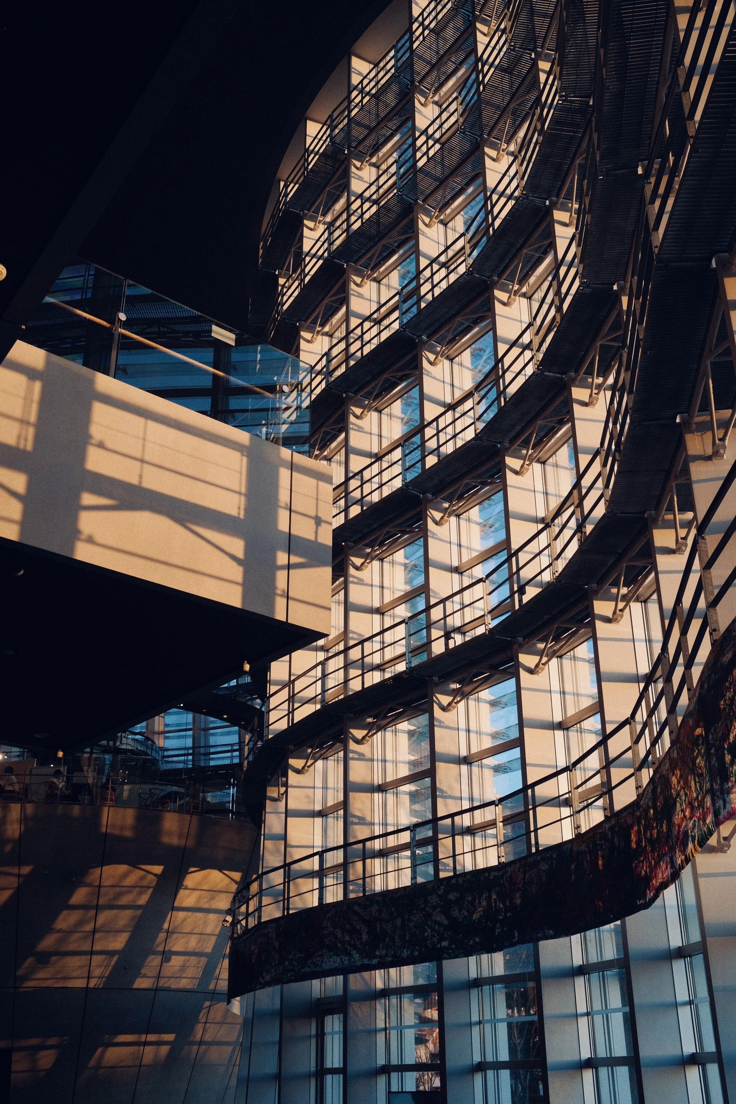 Multiple floors with glass walls and balconies in a modern building, illuminated by warm sunlight, casting shadows.