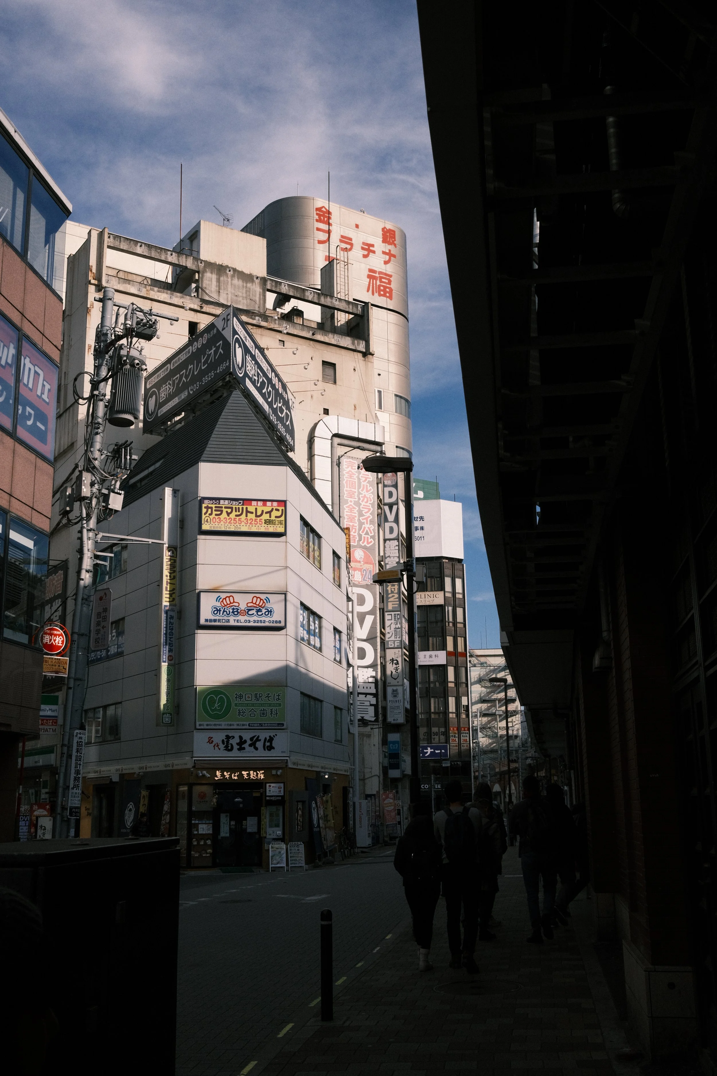 City street view with tall buildings, signage in Japanese, and pedestrians walking on the sidewalk under a partly cloudy sky.