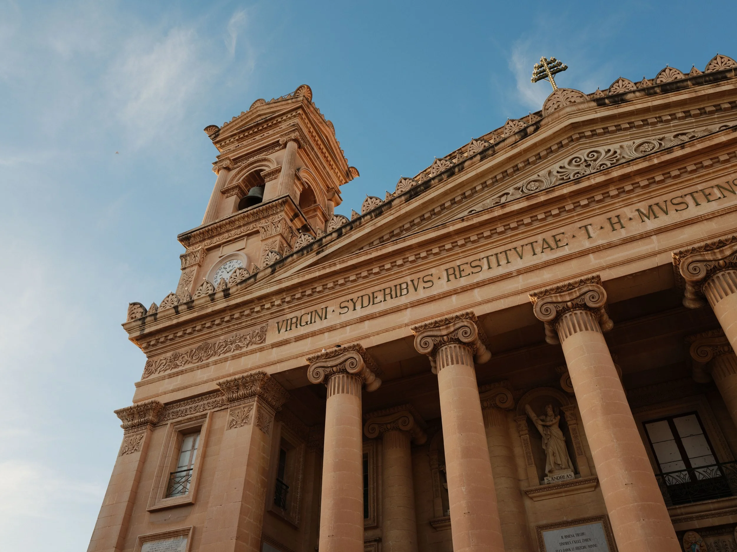 A historic stone building with classical architecture, featuring columns, ornate carvings, a clock, and a cross on top, under a blue sky with some clouds.
