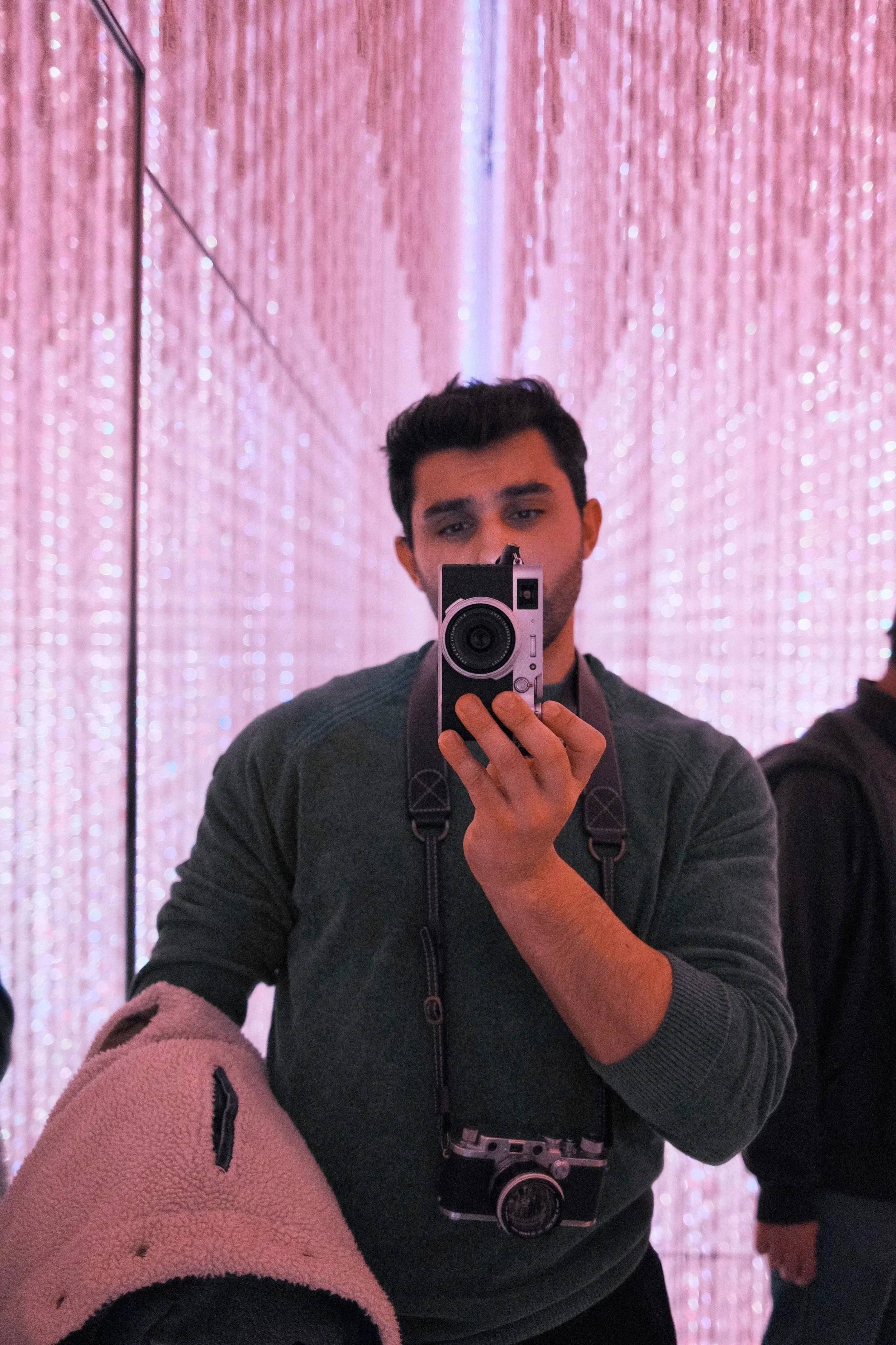 Young man taking a selfie in front of a mirror with pink illuminated walls, holding a camera and wearing a backpack.