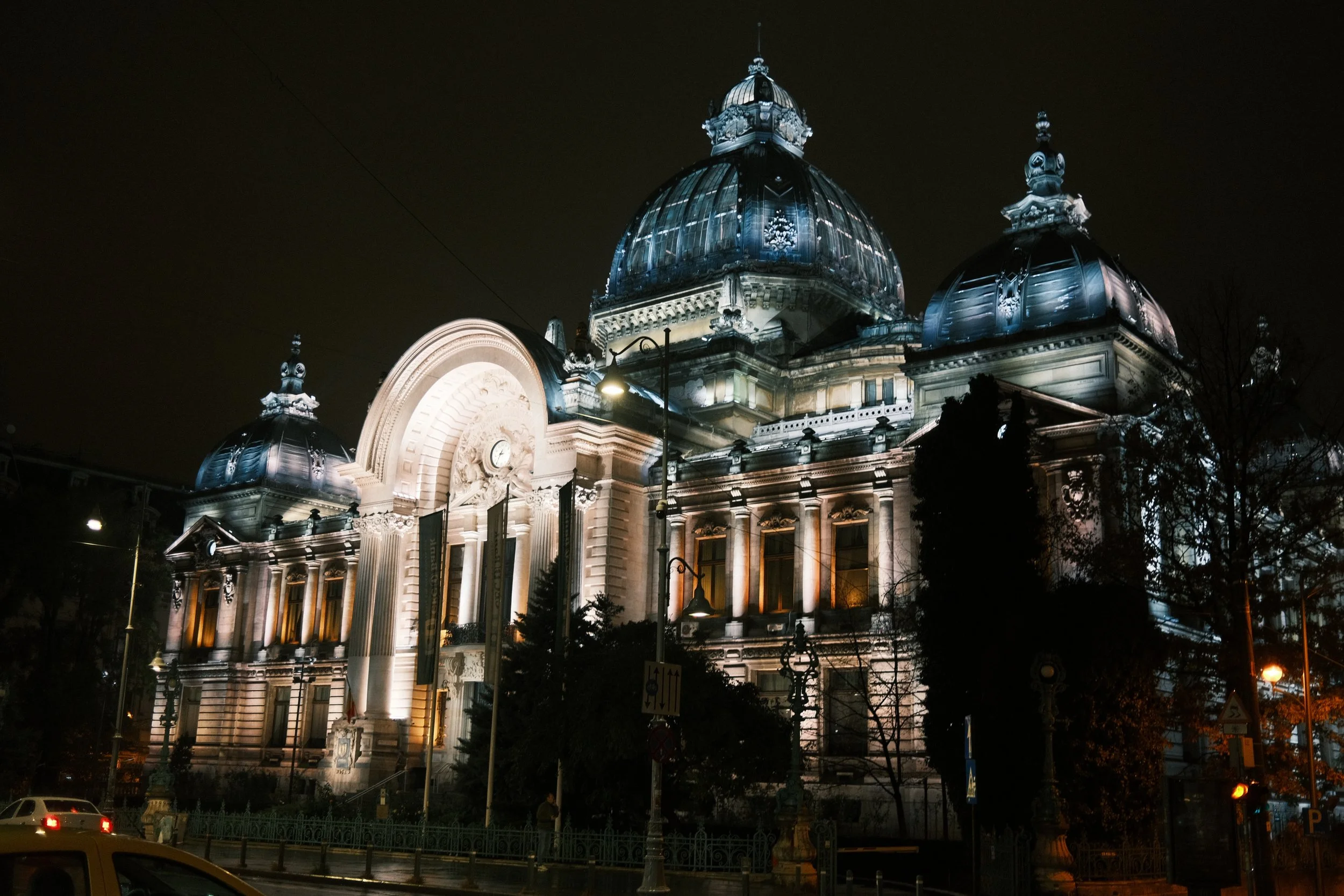 A historic building illuminated at night, featuring large domes and ornate architectural details, with streetlights and trees in the foreground.