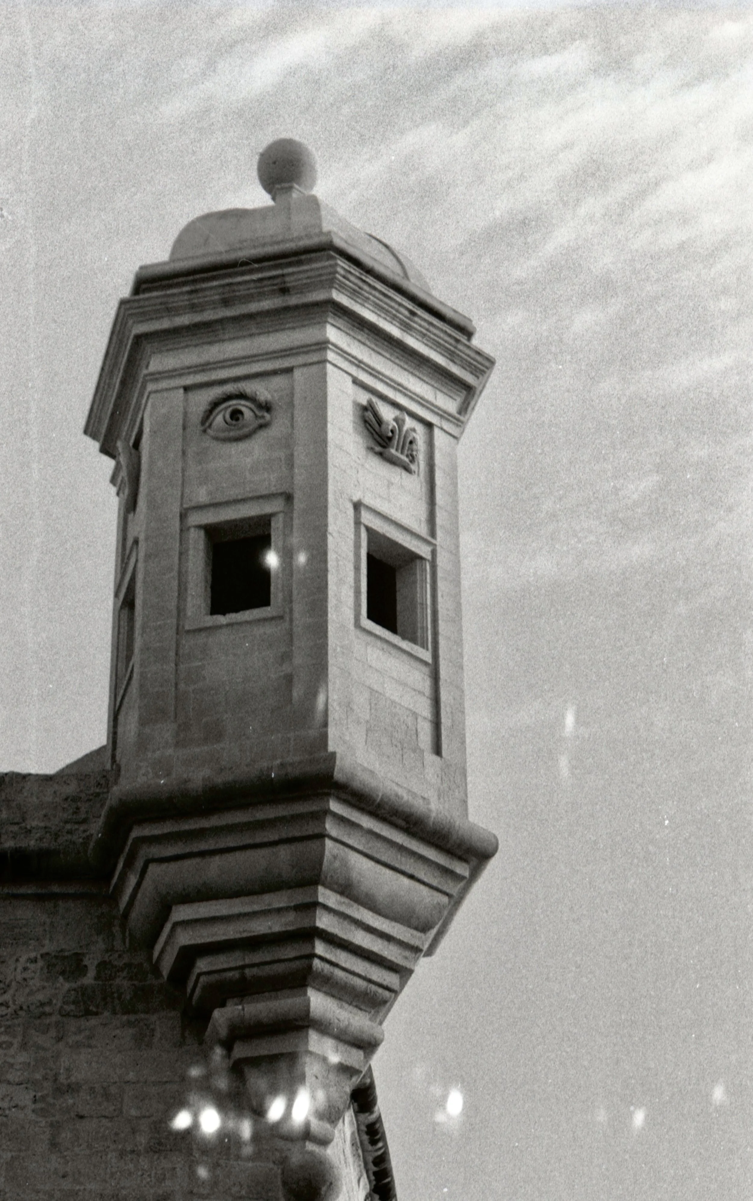 Black and white photograph of a clock tower with decorative eye and bird sculptures near the top, showing a cloudy sky in the background.