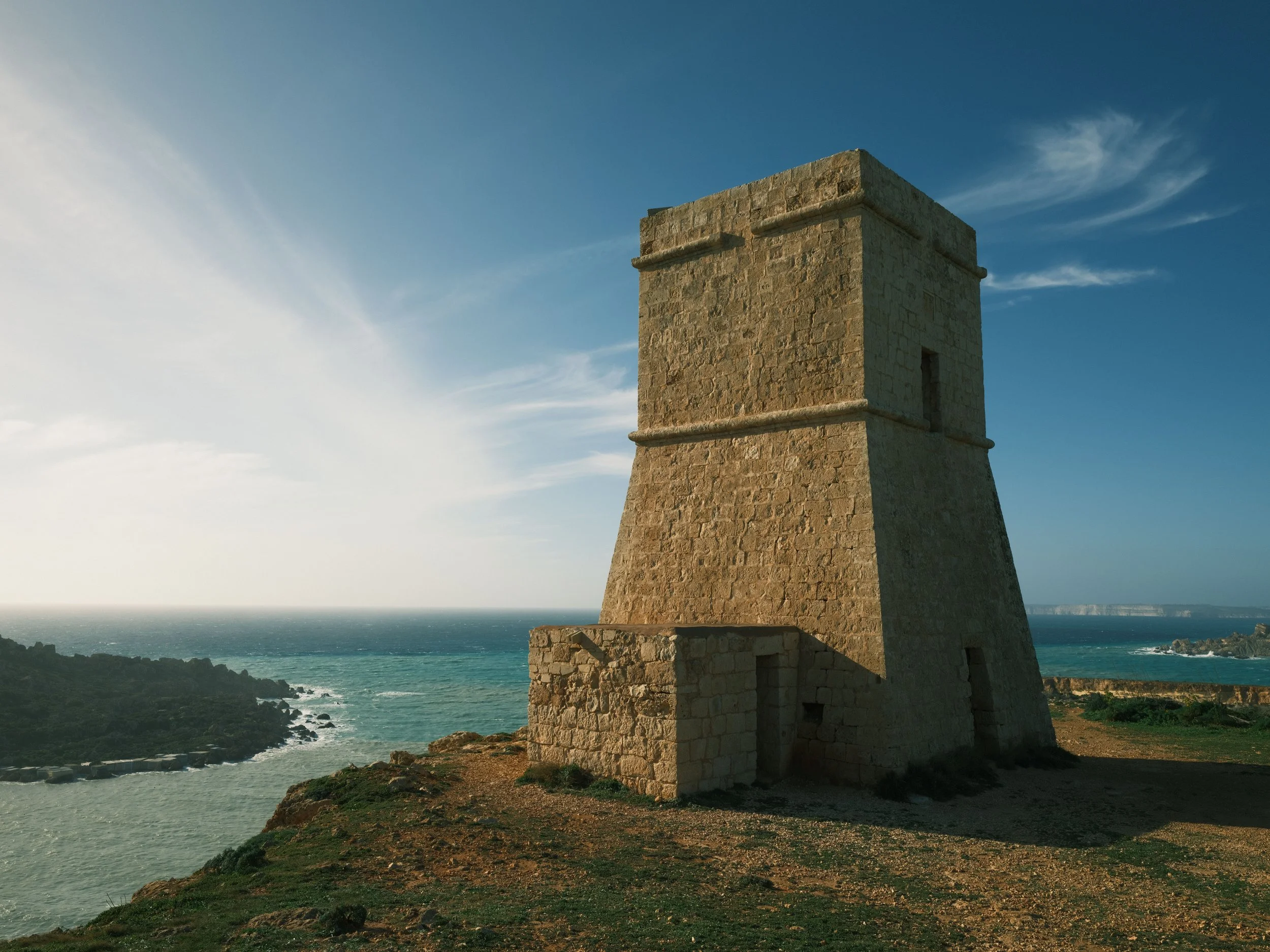 A stone tower with a narrow entrance, situated on a rocky coastline with ocean waves and cliffs in the background under a partly cloudy sky.