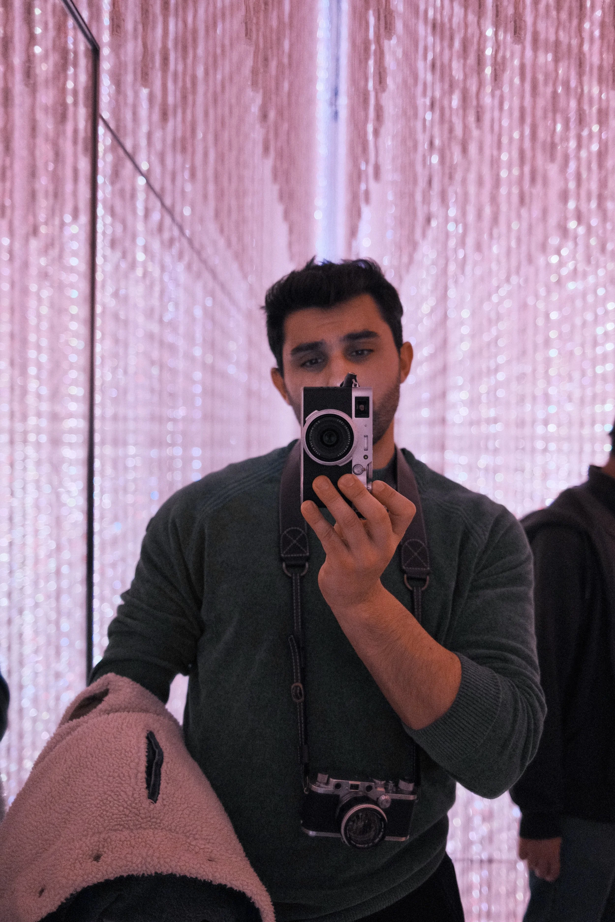 A man taking a selfie in front of a pink, sparkly, curtain-like background. He is holding a camera in front of his face and wearing a dark green sweater with a camera hanging around his neck. Another person is partially visible on his right side.