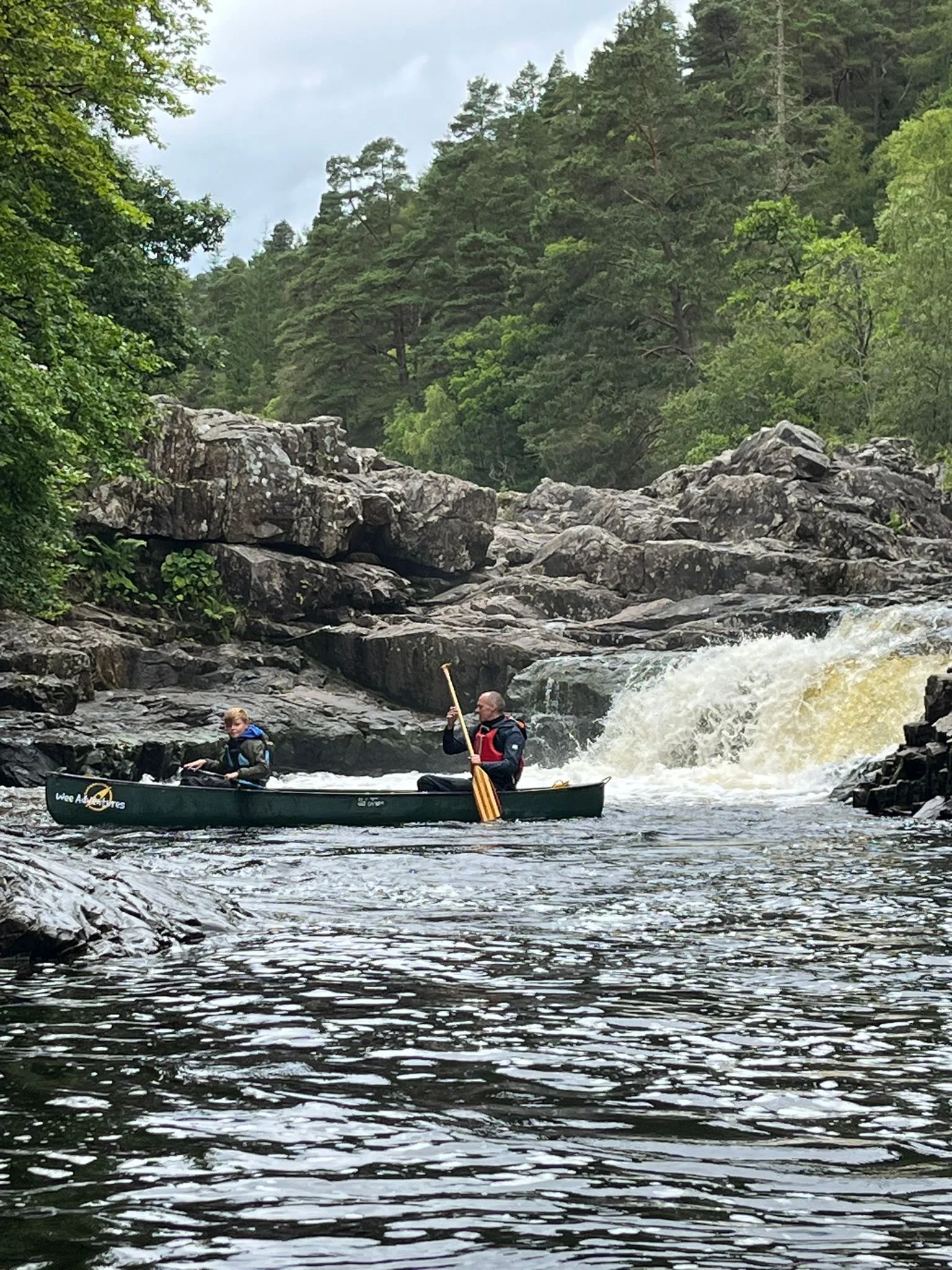 Two people canoeing on a river with rocky banks and surrounding forest scenery.