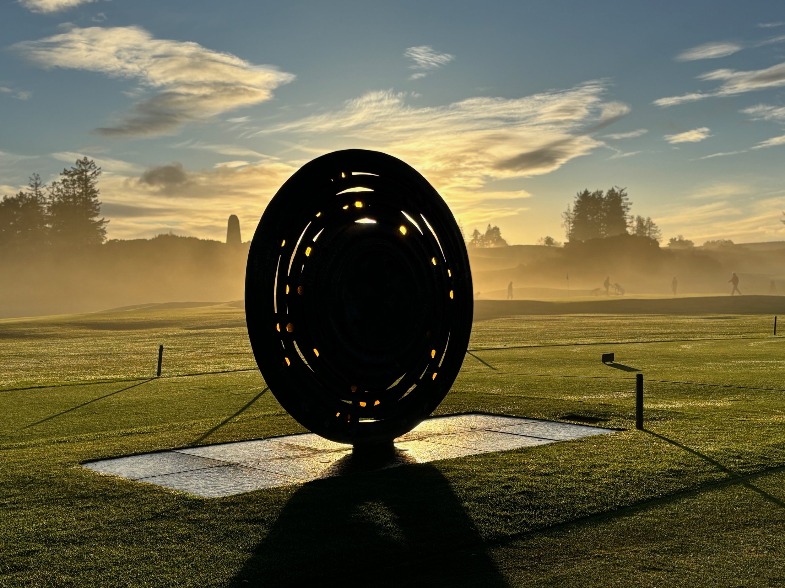 Silhouette of a disc-shaped sculpture on a golf course at sunrise, with mist in the background and trees on the horizon.