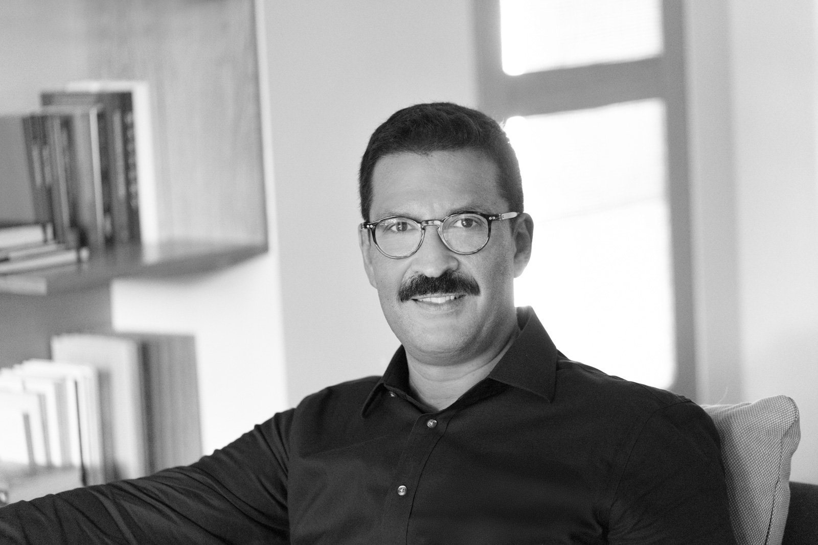 Black and white photo of a man with glasses and a mustache sitting indoors near a window, with bookshelves in the background.