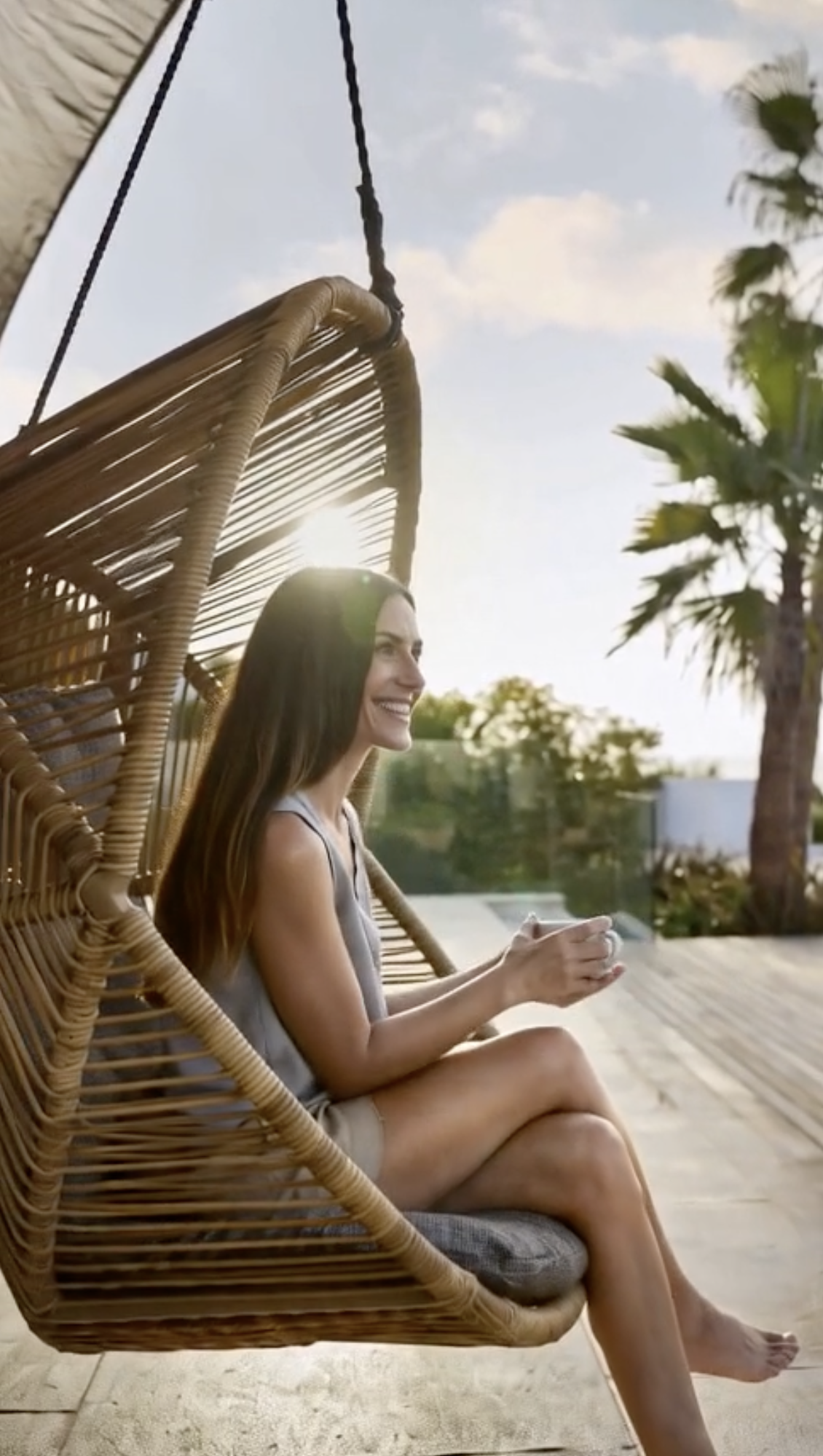A woman sitting in a hanging wicker chair outdoors, smiling and holding a mug, with sunlight and palm trees in the background.