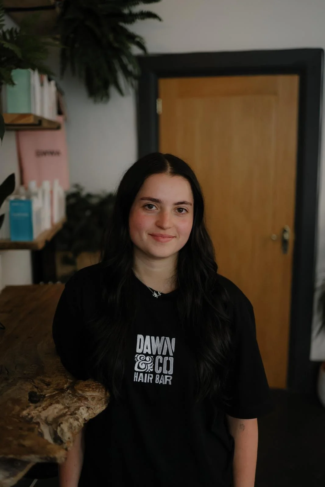 Woman with long curly red hair and glasses smiling in a cozy salon or coffee shop, wearing a black hoodie that says 'Dawn' with a design, standing near a table with red brochures and a mirror.