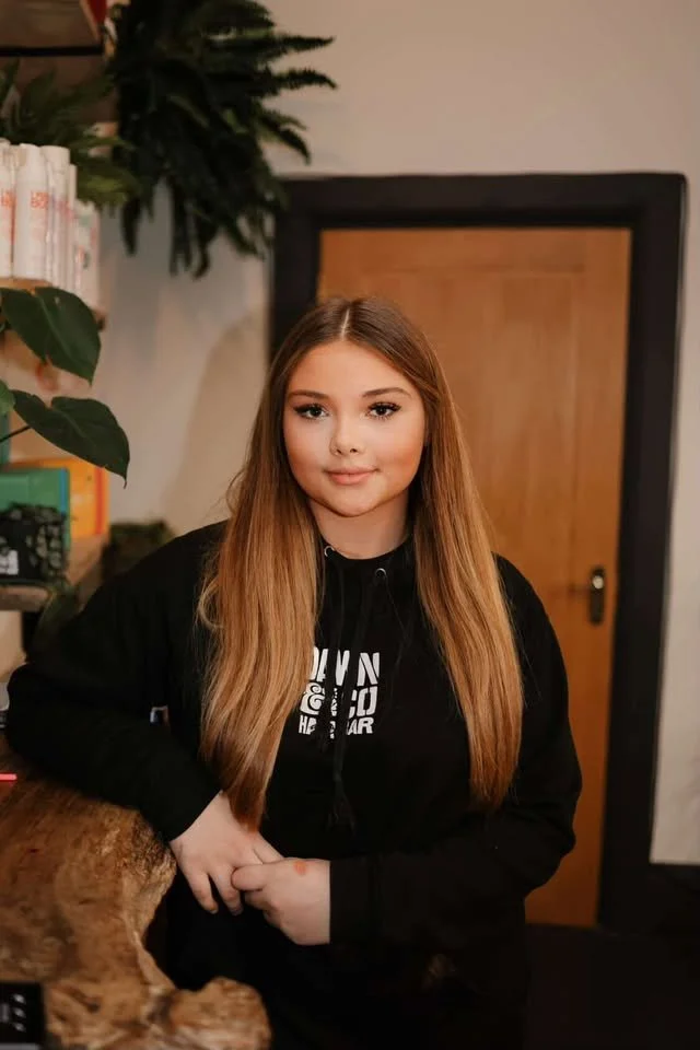 A young woman with long, straight, light brown hair, wearing a black hoodie, standing indoors near a wooden door, with her left hand resting on a wooden surface, smiling gently at the camera.