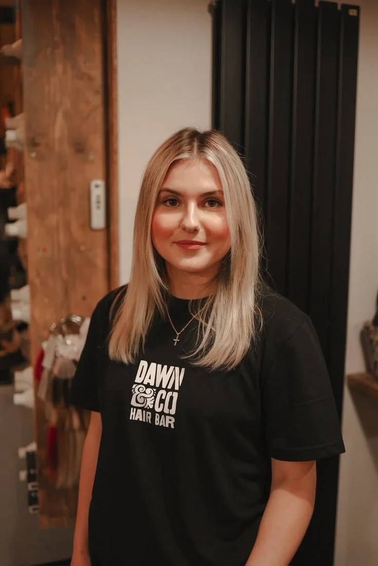 Young woman with red hair wearing a black hoodie, standing near indoor plants, wooden door in background.