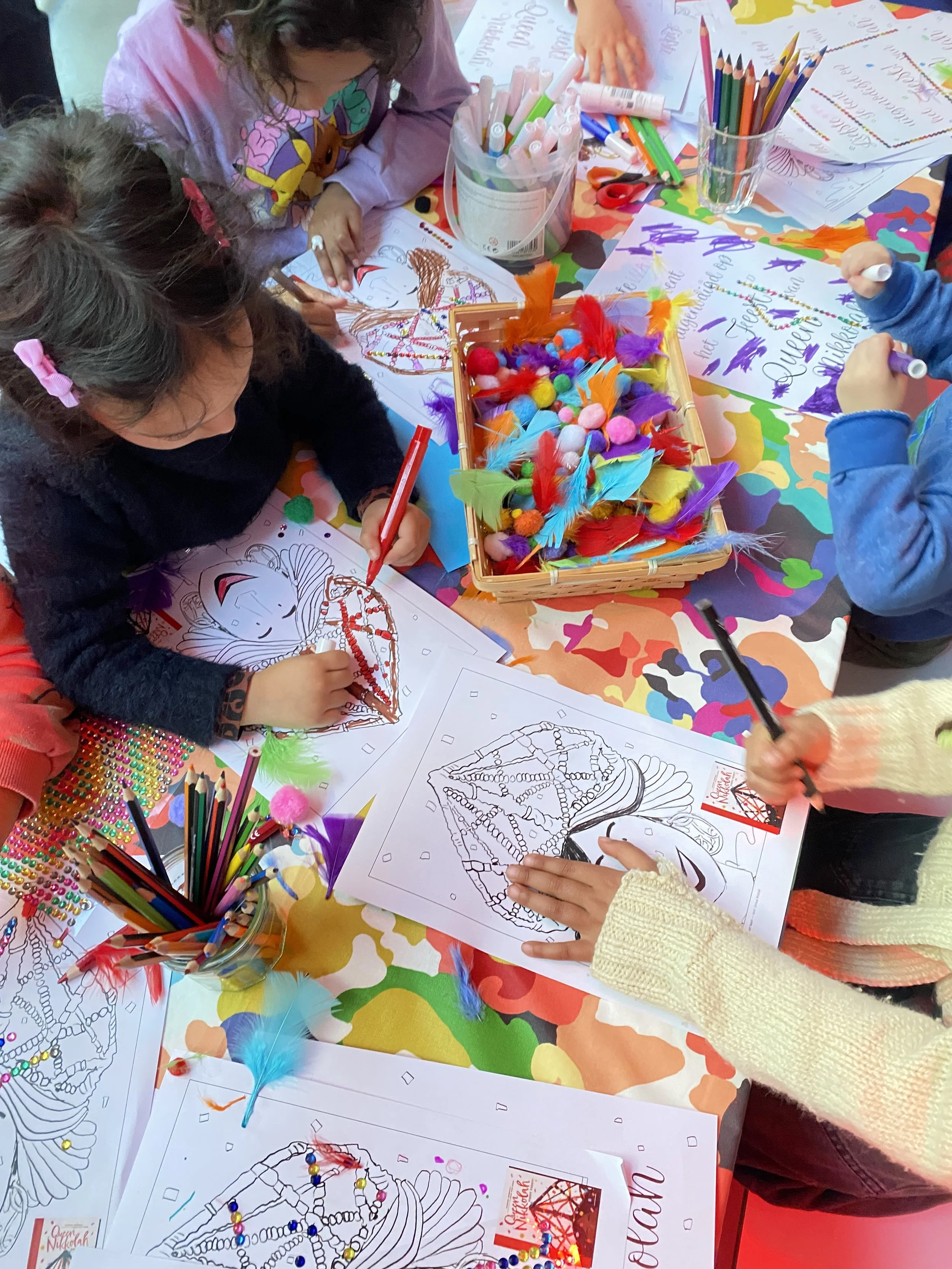 Kinderen kleuren herfstblaadjes op een tafel met tekenpapier en kleurrijke materialen zoals veren, stippen, en glaasjes met kleurpotloden.