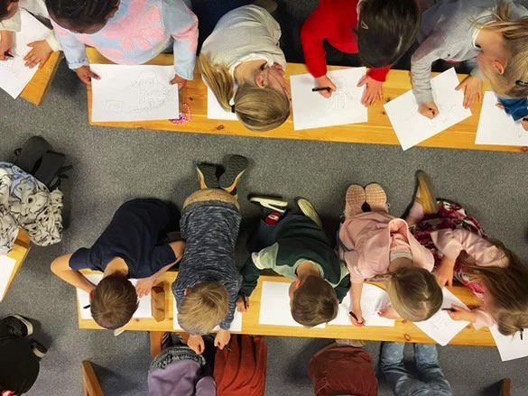 Kinderen zitten en liggen op de vloer, tekenen en schrijven op papier. Vreemd perspectief van bovenaf.