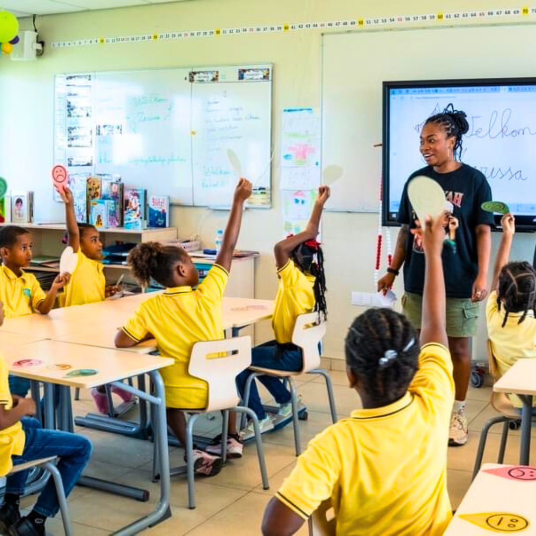 Een klaslokaal met jonge studenten in gele uniformen, enkele leerlingen steken hun hand op terwijl een lerares bij de whiteboard staat.