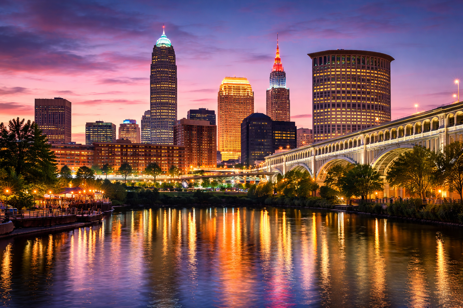 Nighttime view of the Cleveland skyline, with illuminated downtown buildings under a purple and pink dusk sky. A lit bridge curves in from the right foreground, and warm city lights glow across the scene.