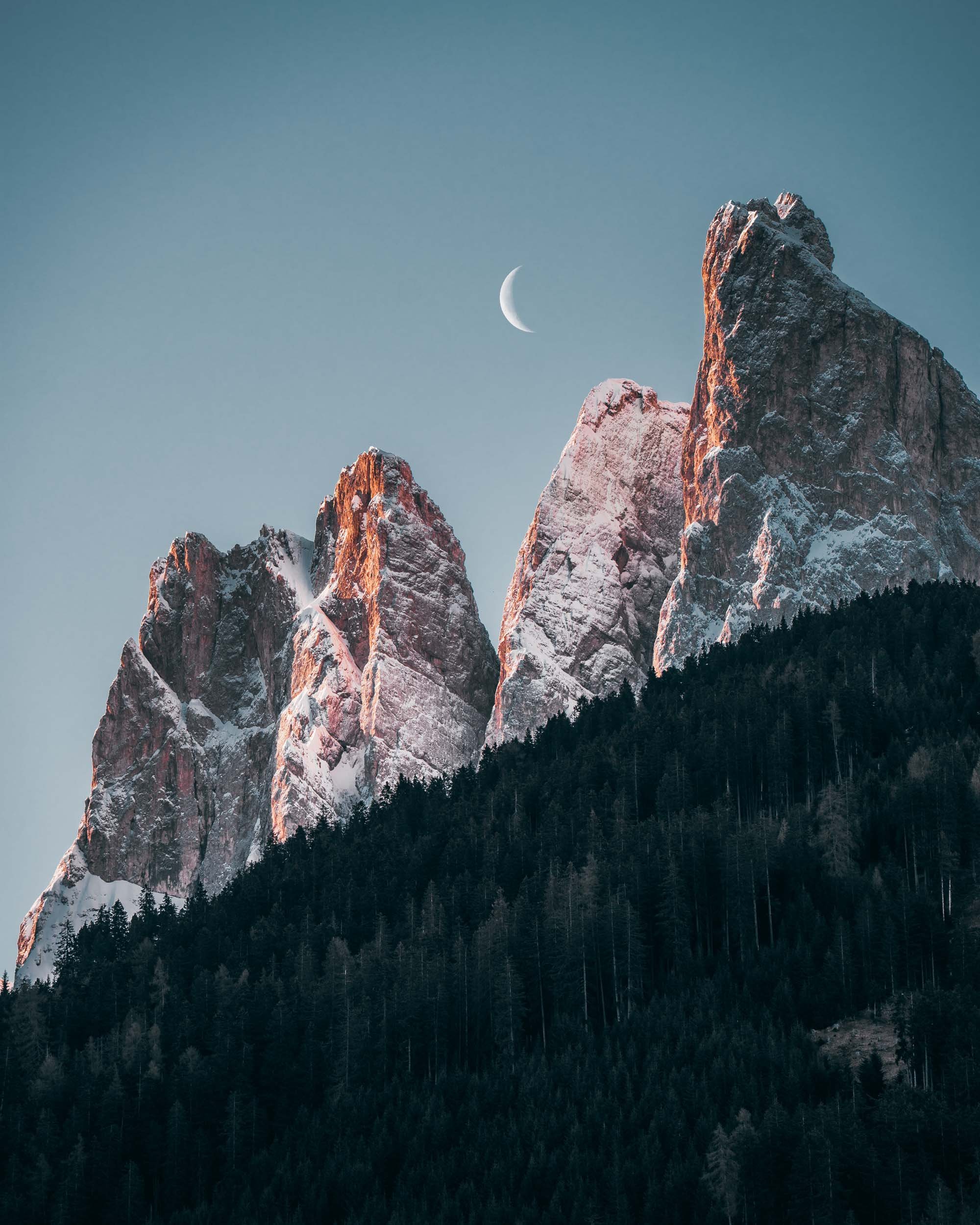Snow-capped mountain peaks with a crescent moon in the sky above a forested landscape.