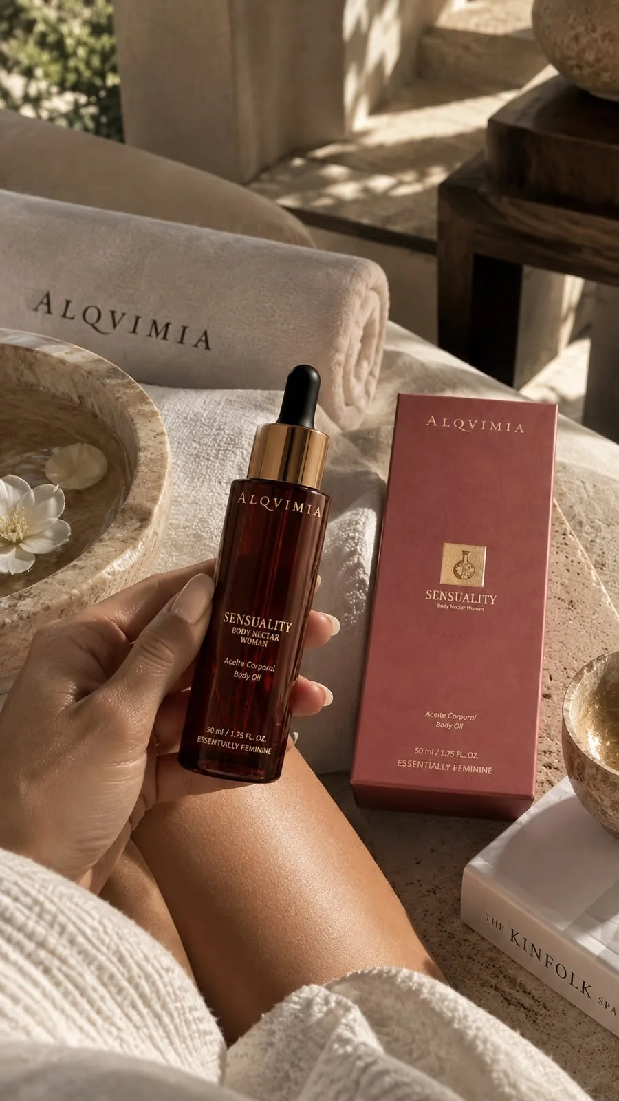 A bottle of Alqvimia Sensuality Body Nectar Woman body oil held in a person's hand, with a matching pink box beside it. The setting includes a white towel with the word Alqvimia embroidered, a bowl with floating white flowers, and a cozy, sunlit outdoor or near-window space with a beige and natural decor theme.