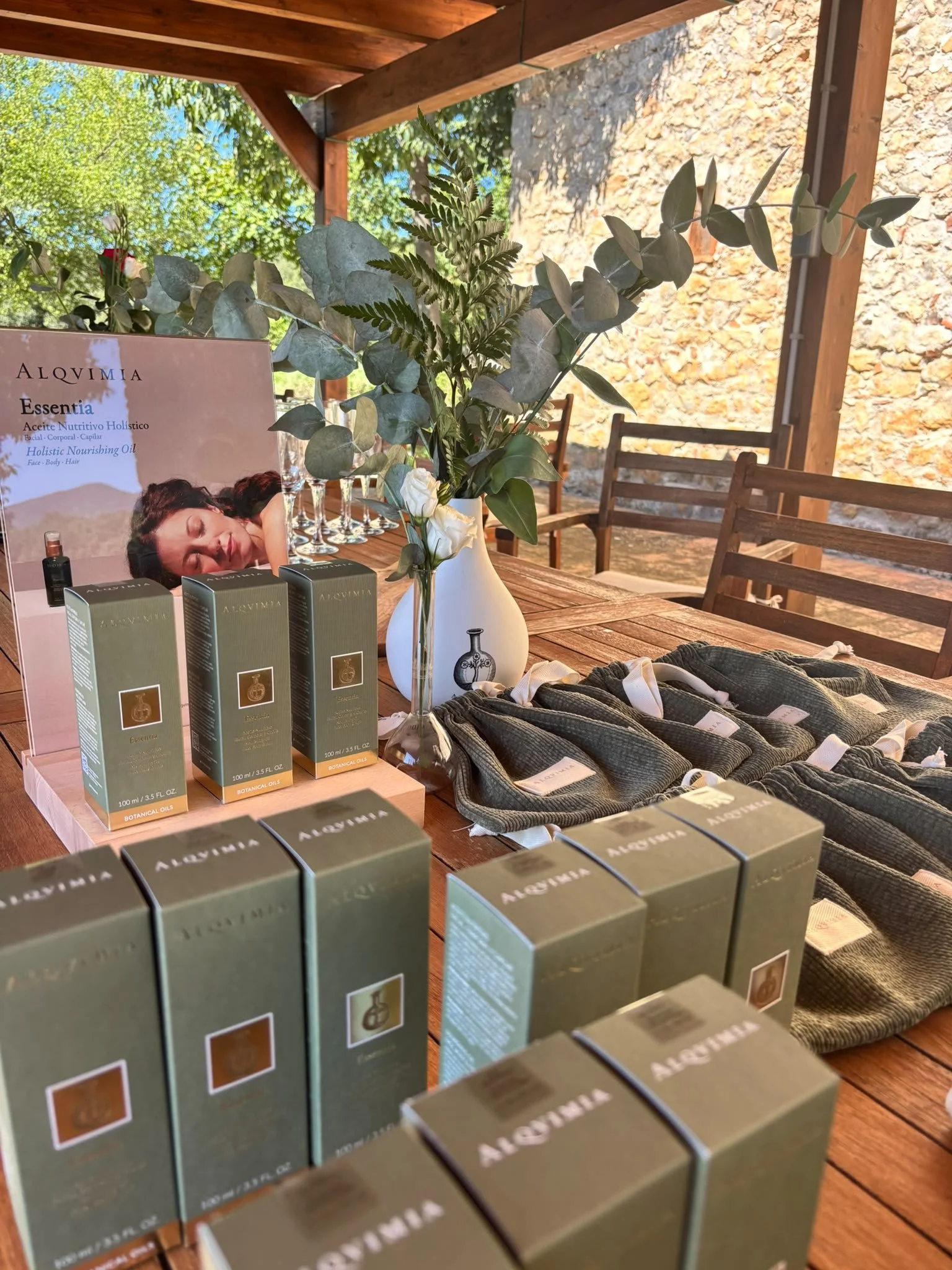 Display of Alquimia skincare products including boxed items and small bottles on a wooden table, decorated with a vase of mixed green and white flowers, outdoors under a wooden shelter with a stone wall and trees in the background.