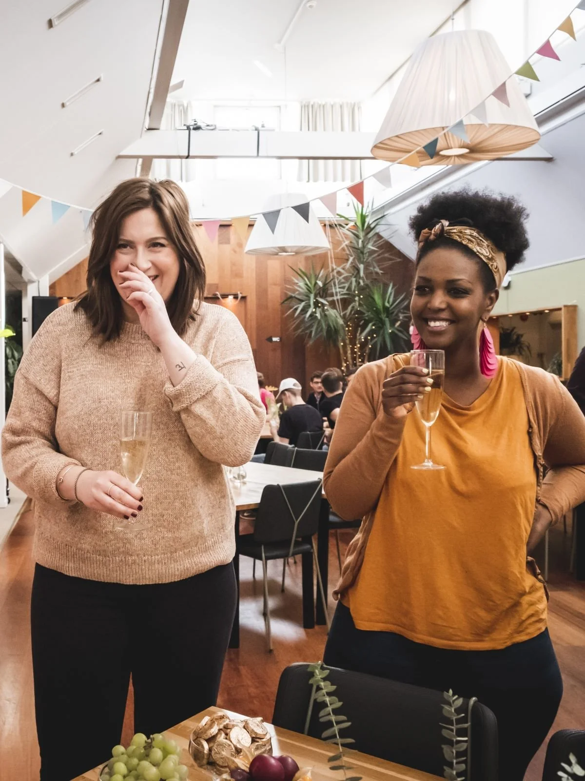 Två kvinnor som andas och skrattar, båda håller glas med champagne, i ett festligt inomhusarrangemang med bunting och växter på Teamfabriken.