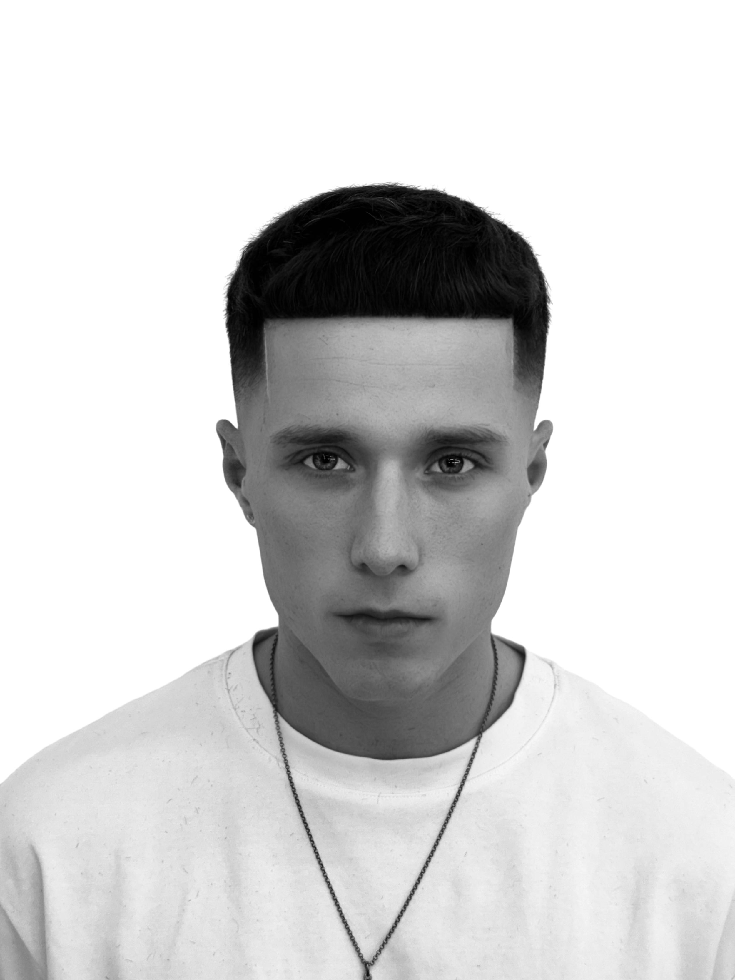 Black and white photo of a young man with short dark hair and piercing eyes, wearing a plain white t-shirt and a necklace, facing forward against a black background.