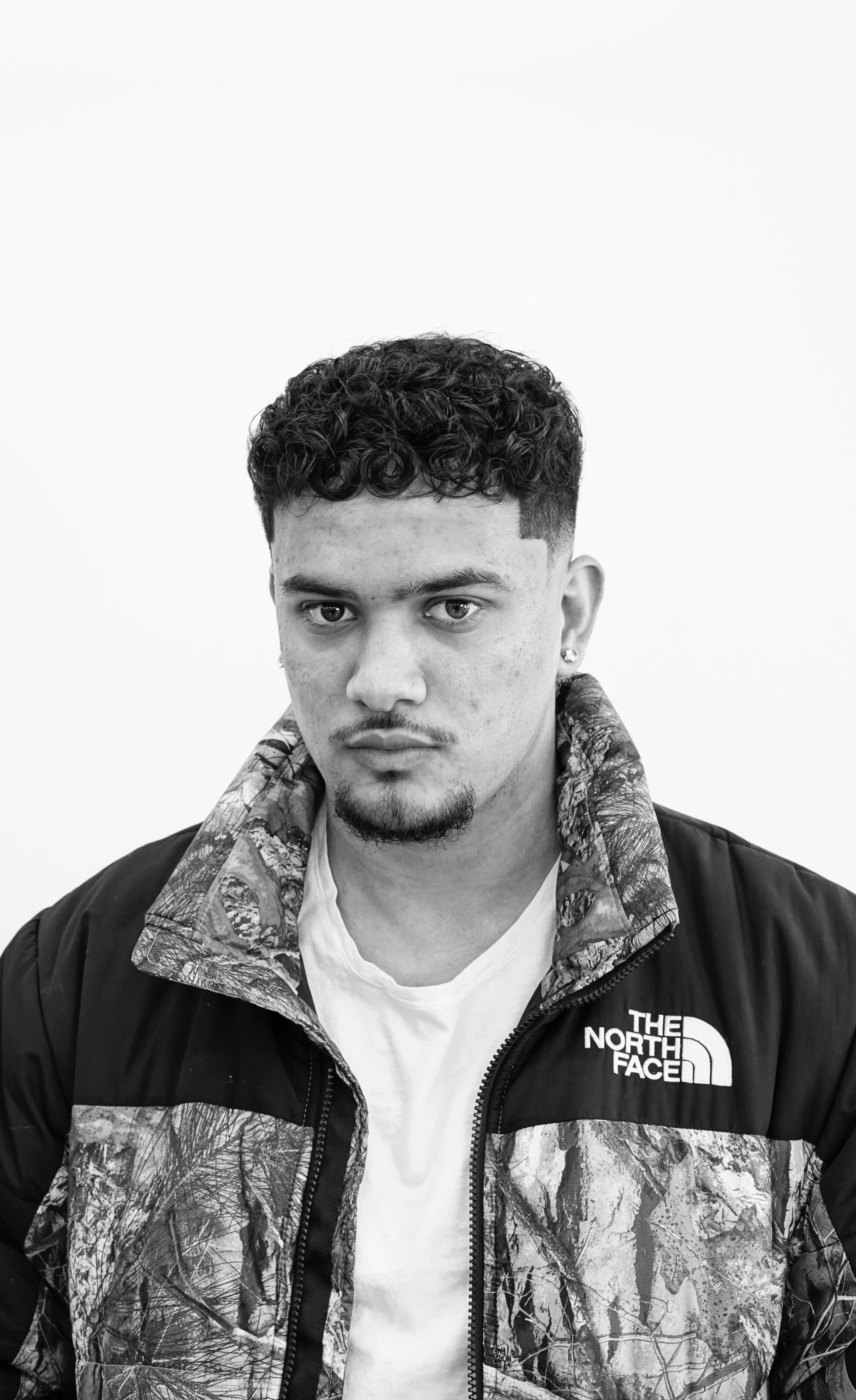 Black and white portrait of a young man with curly hair and a goatee, wearing a camouflage patterned The North Face jacket over a white T-shirt, looking directly at the camera against a plain background.