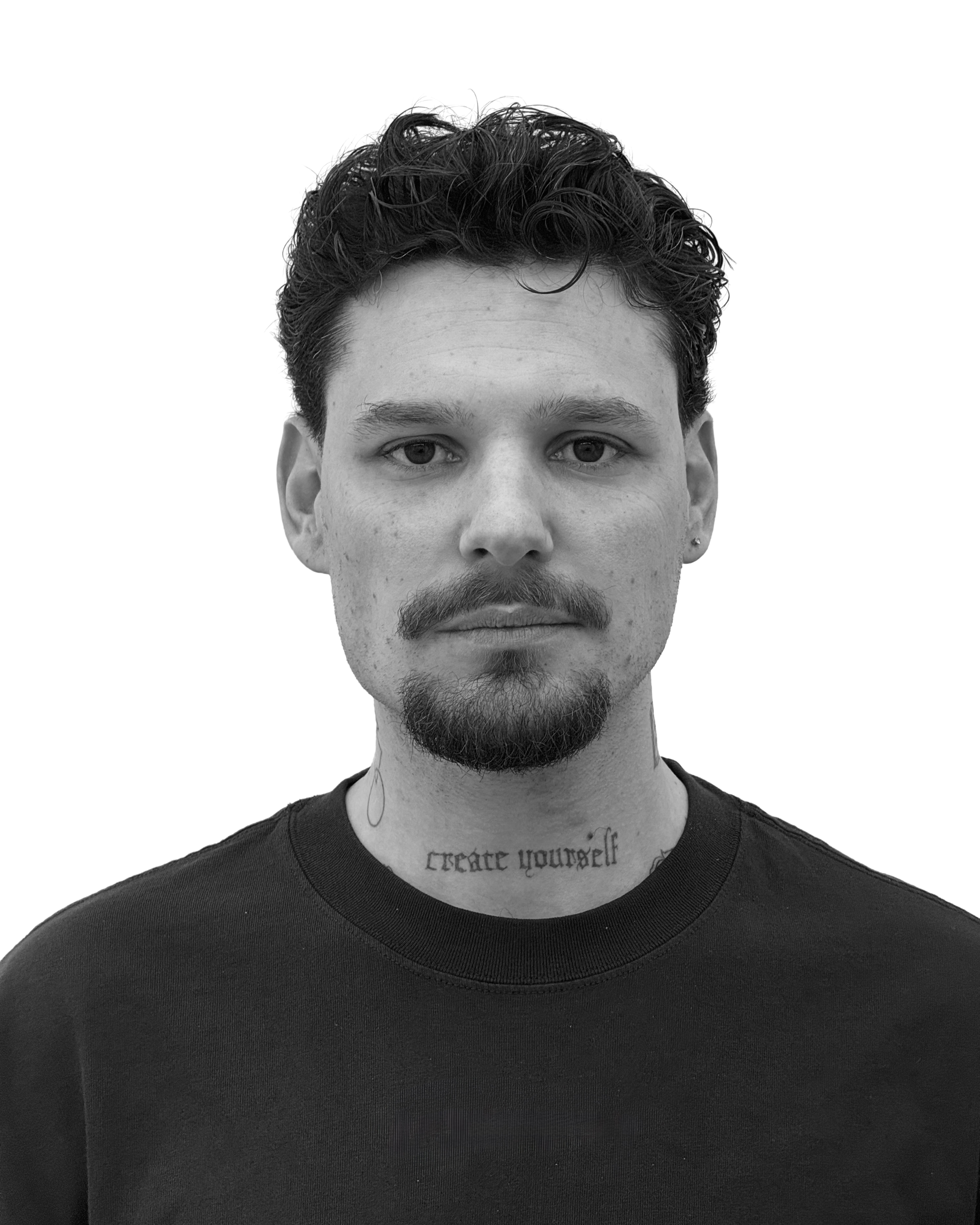 Black and white portrait of a young man with wavy hair, a mustache, a small goatee, and a tattoo on his neck that reads 'create yourself'.
