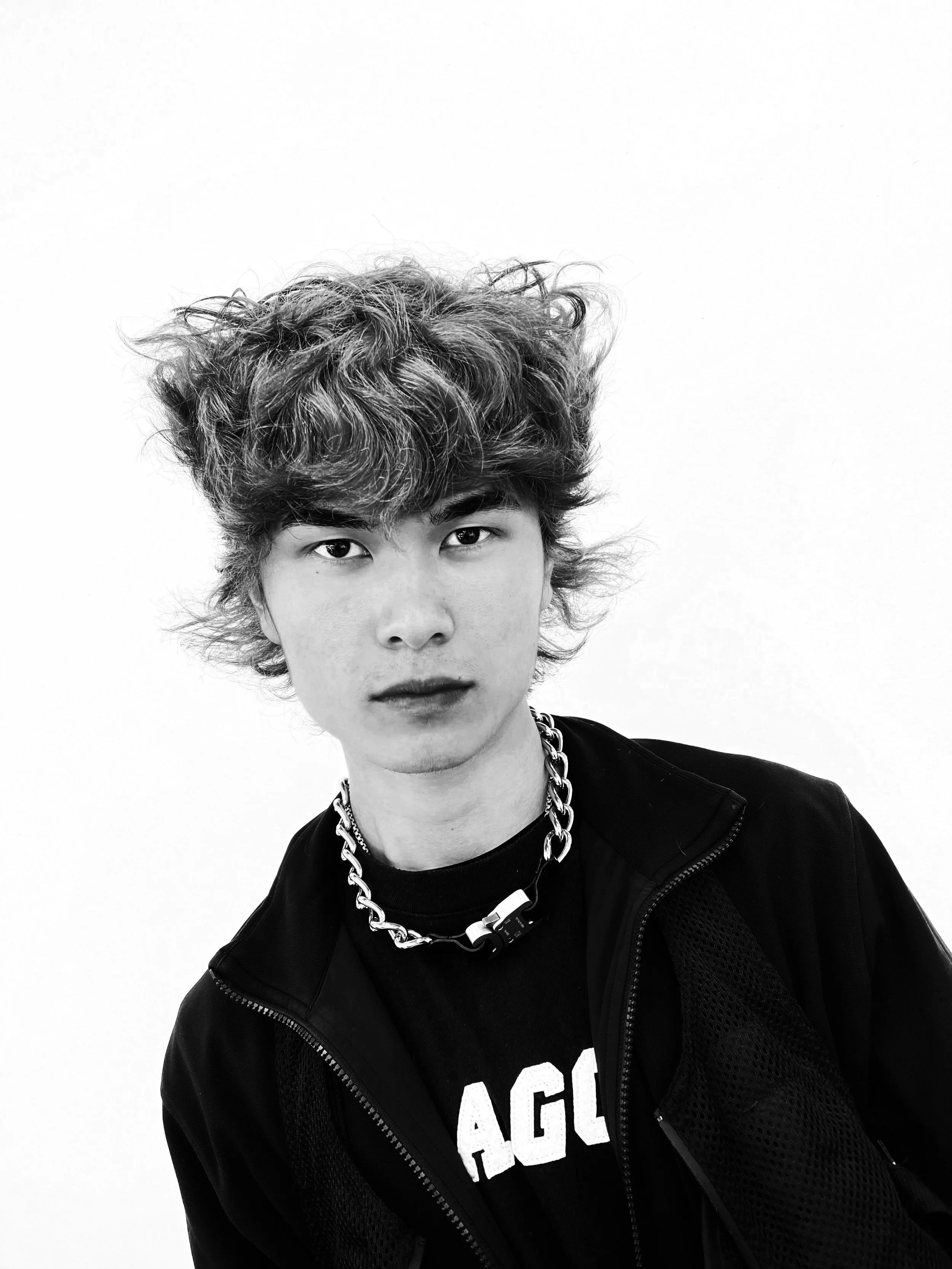 Black and white portrait of a young man with wavy hair, wearing a chain necklace and a zip-up jacket, standing against a plain background.