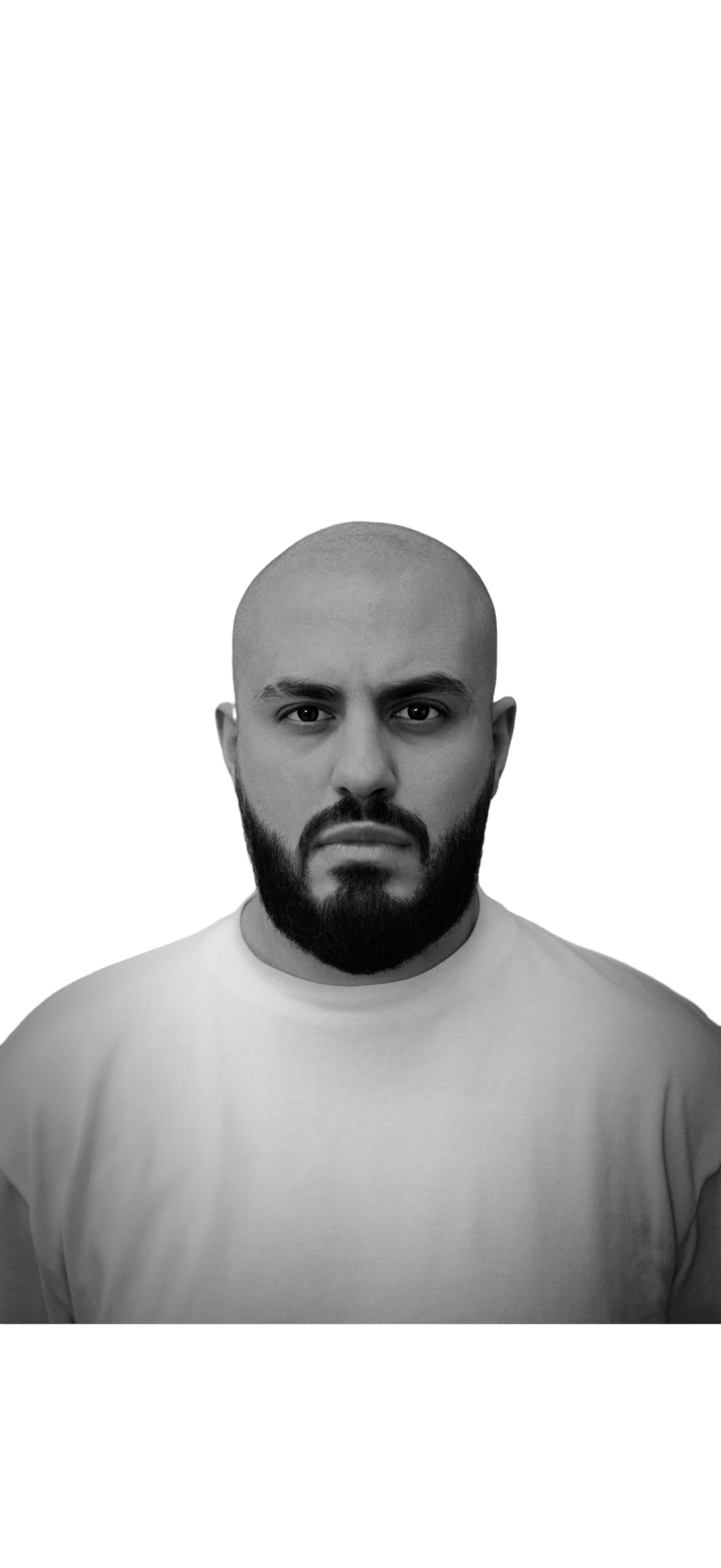 A black and white portrait of a young man with a beard and mustache, wearing a plain light-colored t-shirt, looking directly at the camera with a serious expression.