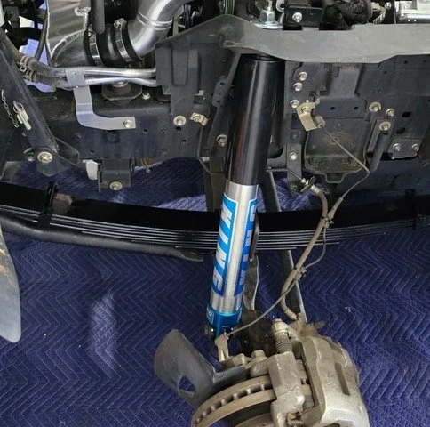 Close-up of a vehicle's suspension system, showing a blue shock absorber and brake caliper, with various mechanical parts in the background.