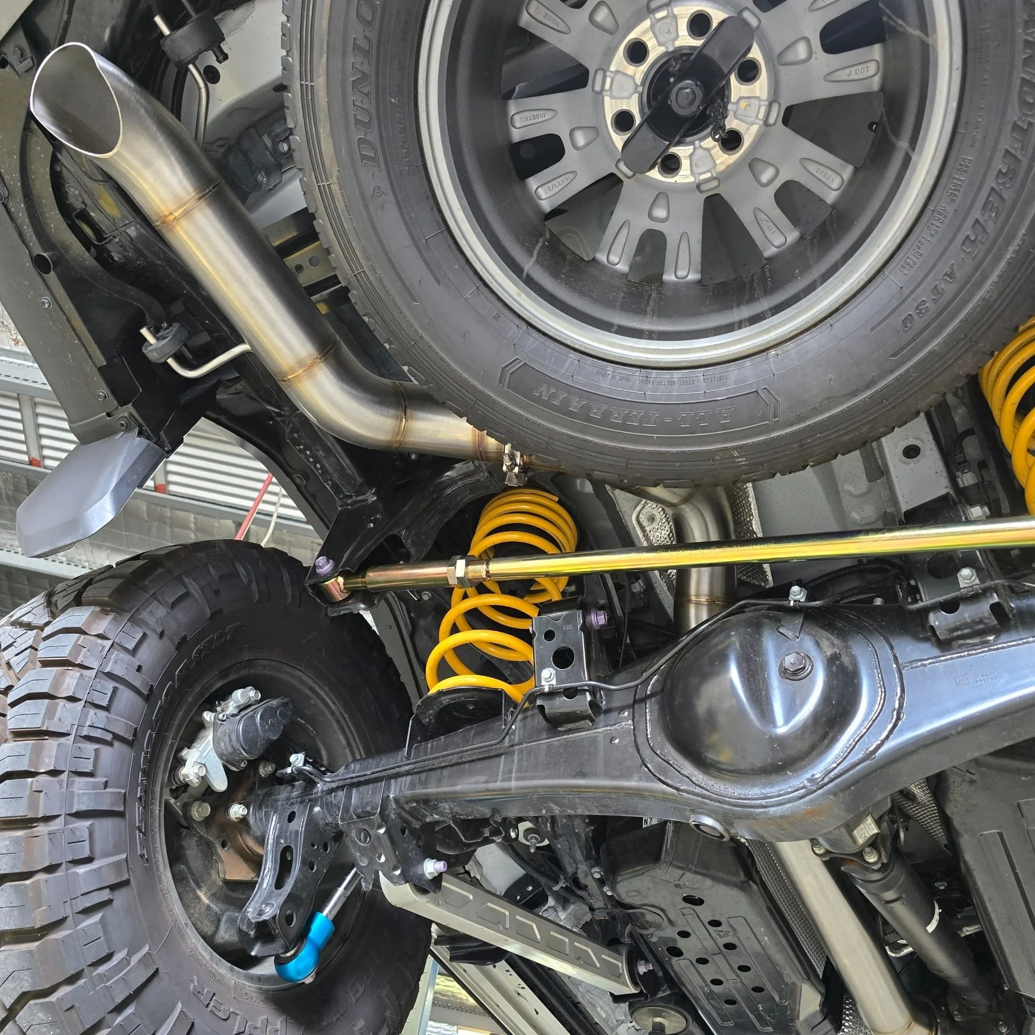 Under an off-road vehicle, showing a rear tire, yellow suspension coil springs, and parts of the exhaust system.