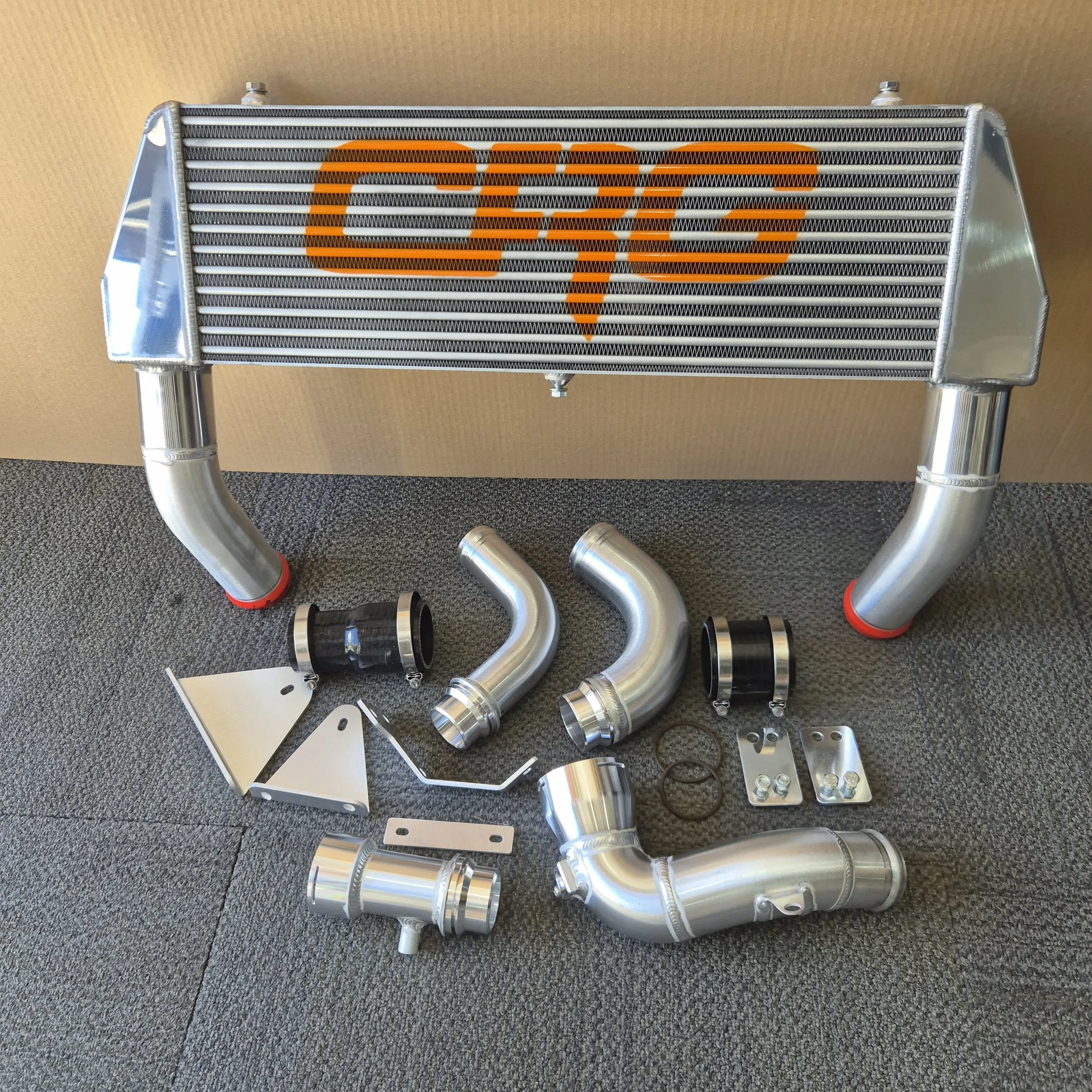 Aluminum car intercooler with orange branding, along with various silver pipes, couplings, and mounting brackets for installation.